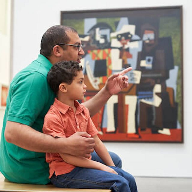 An adult and child sitting on a bench looking and pointing at a painting with the Three Musicians by Pablo Picasso in the background.