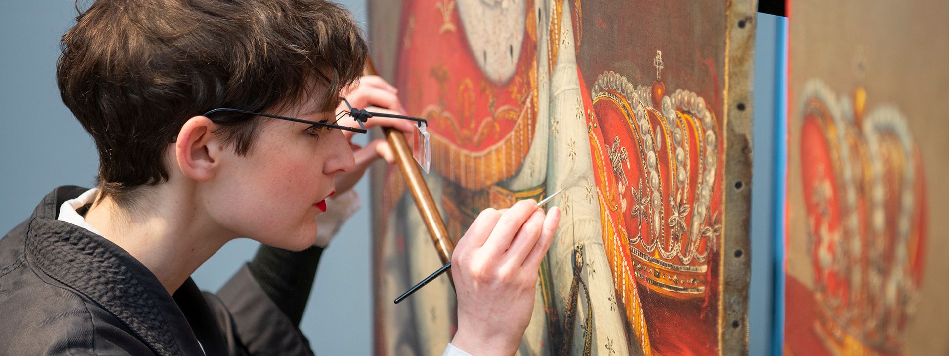 A conservator inspecting a painting with a fine brush and magnifying glass