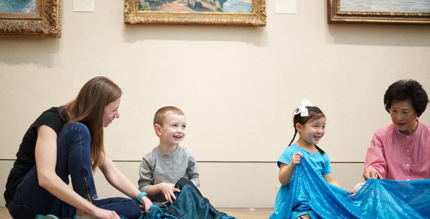 Two children and two adults play with sparkly, blue fabric on the floor of a gallery