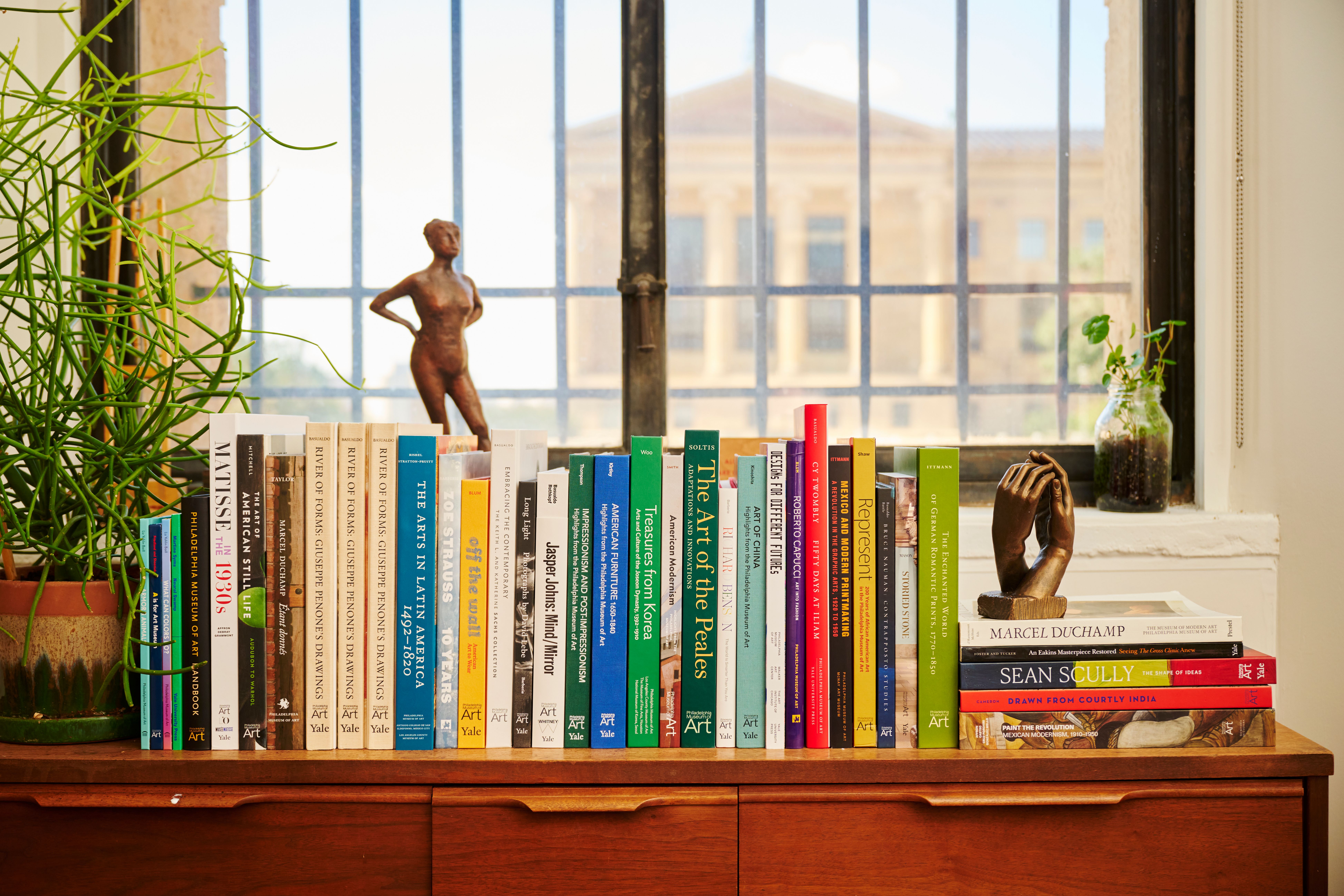 A bookshelf of museum publications and plants by a window
