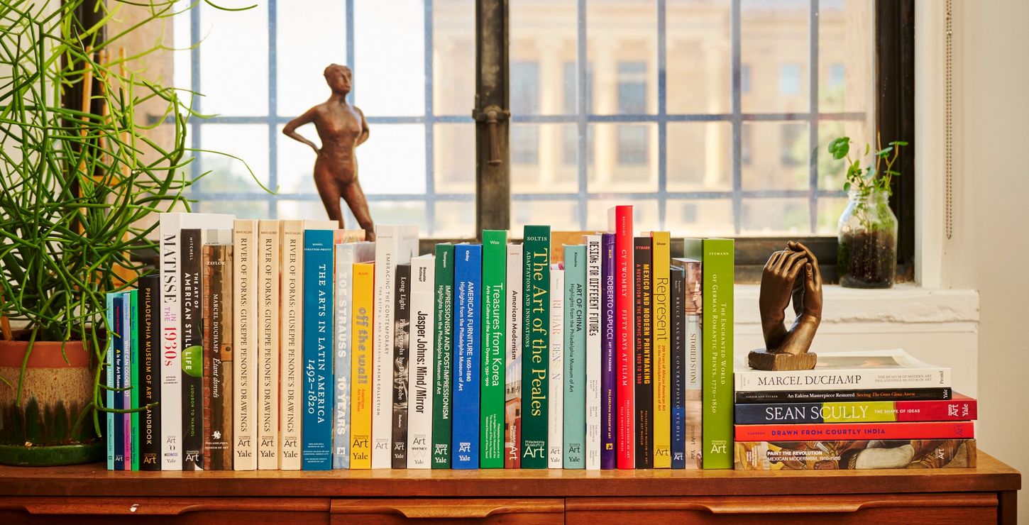 A bookshelf of museum publications and plants by a window