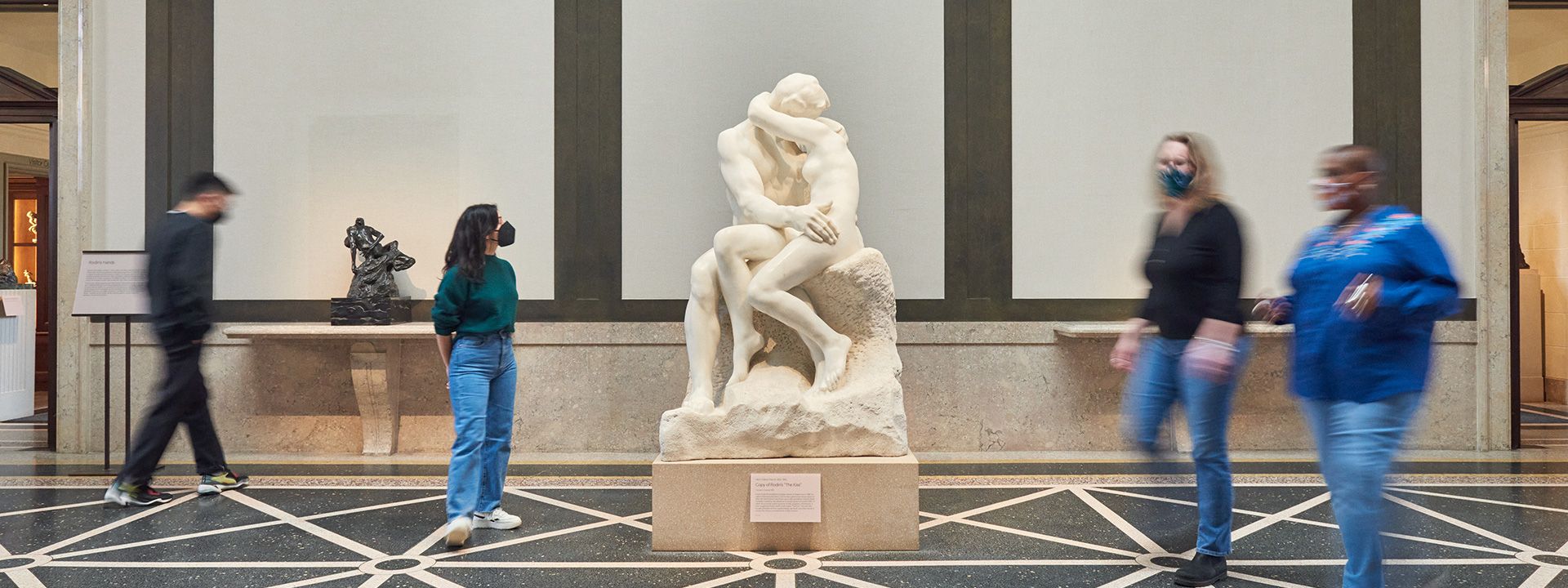 Four people wearing masks walking inside a gallery near a statue
