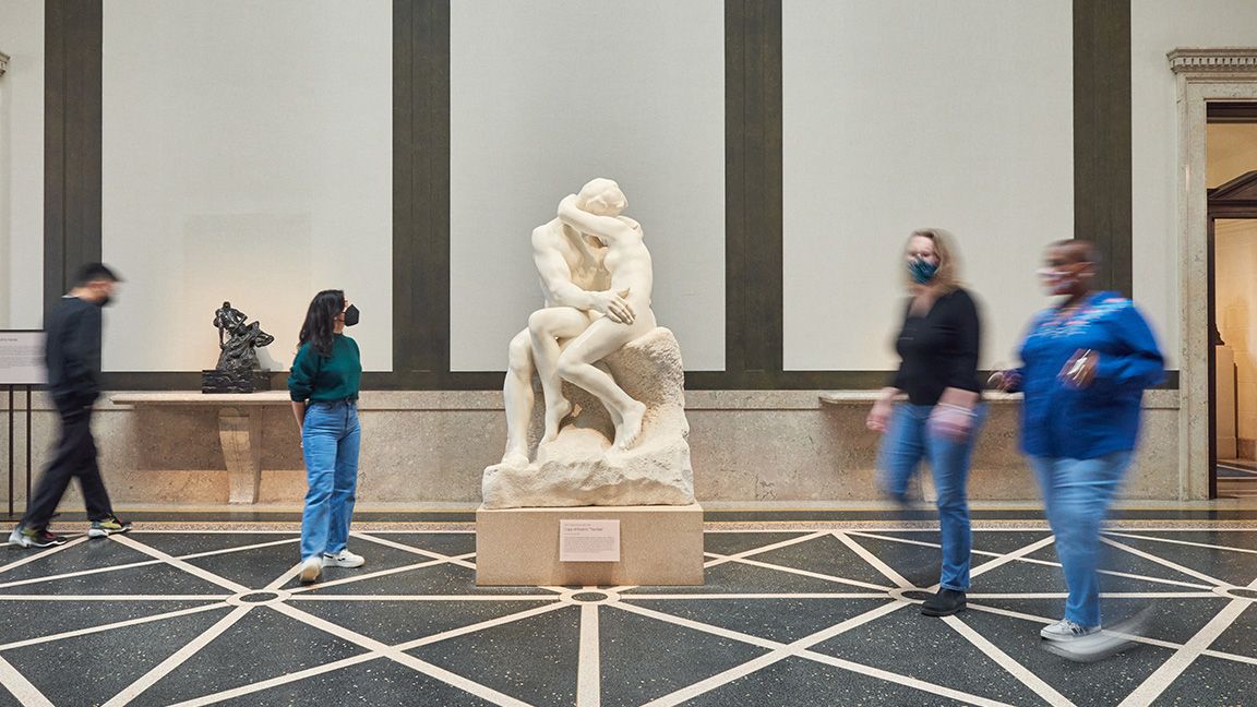Four people wearing masks walking inside a gallery near a sculpture