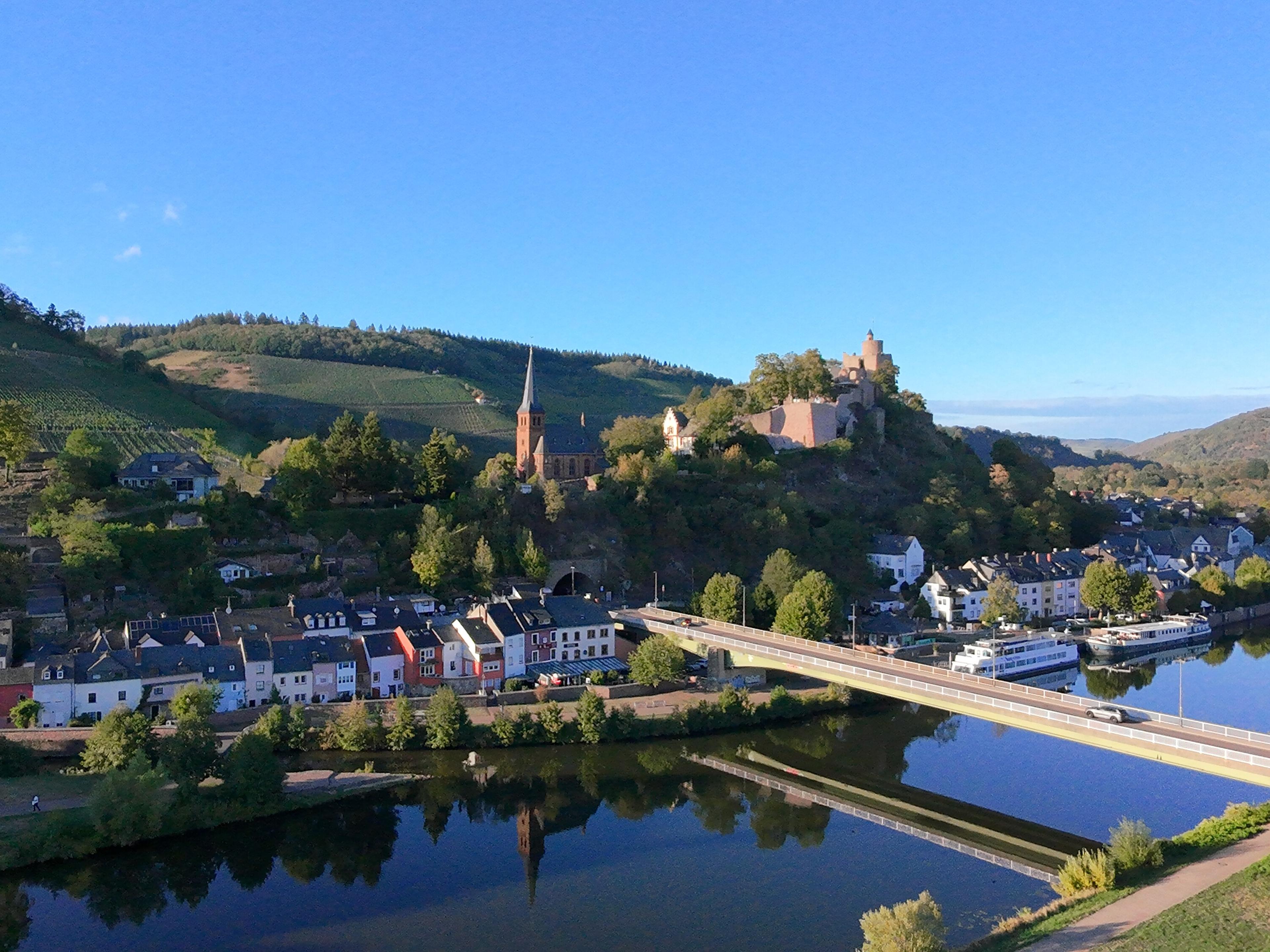 Luftaufnahme von Saarburg: bunte Häuser am Saarufer, die Brücke über die Saar, darüber ein Kirchturm und die Burg vor Weinbergen