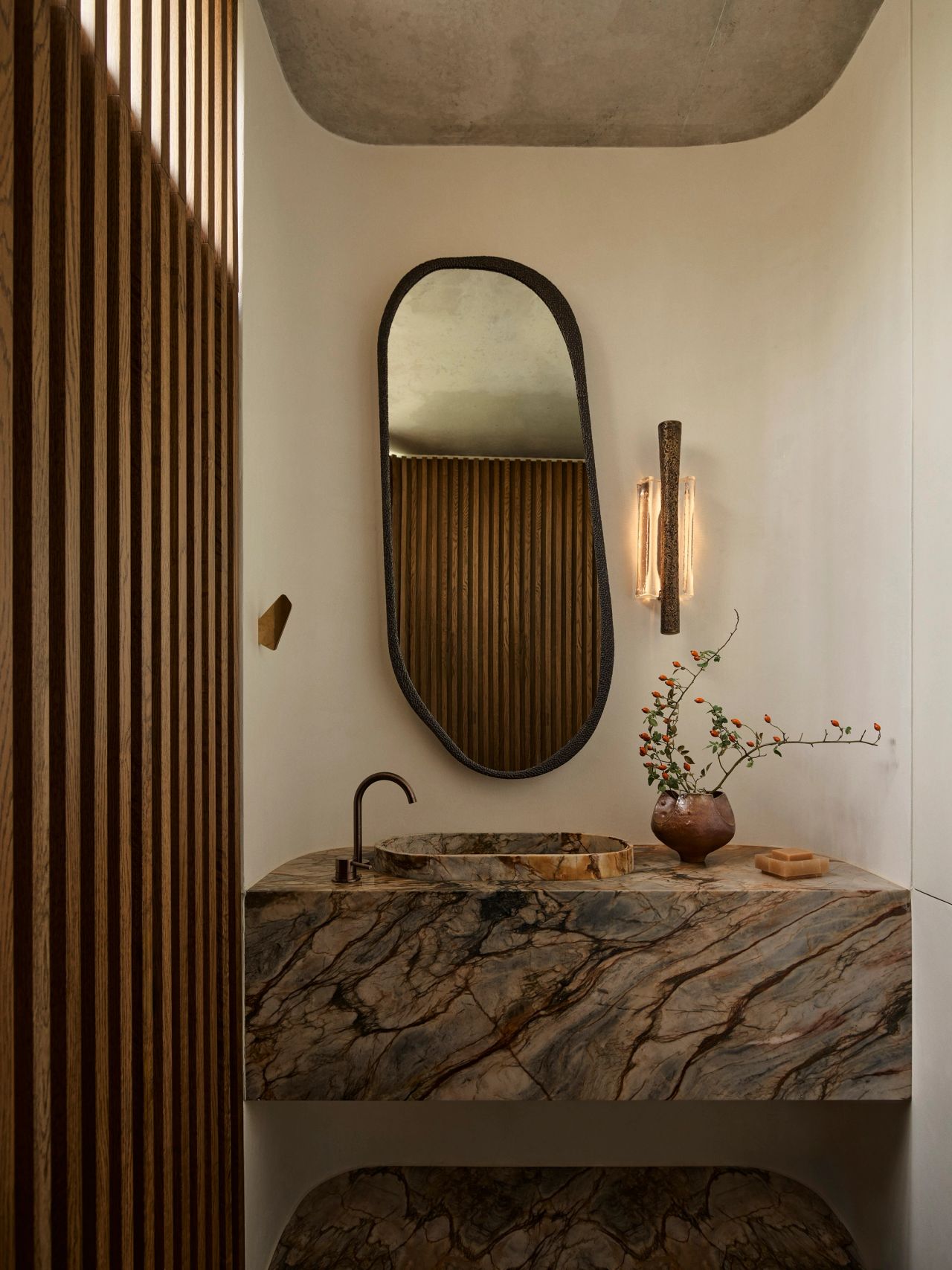 Orla powder room with a curved blue, cream, and rust-veined stone vanity, aged brass hardware, and an irregular oval mirror in a brass frame. A bronze and glass cylindrical wall light hangs beside it, and a brown-rust ceramic vase with sculptural florals sits on the vanity. Timber batten wall details are reflected in the mirror.