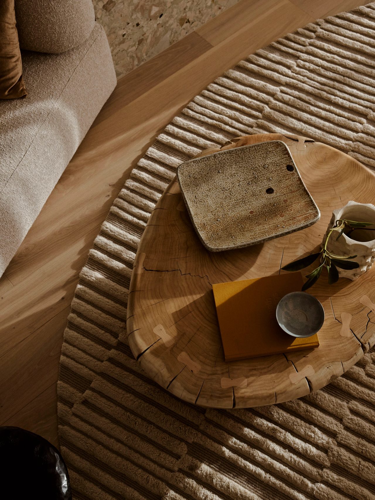 Patchwork Bird’s-eye view of a solid timber coffee table styled with decorative ceramics, positioned over a beige striped oval rug. A beige boucle sofa is partially visible in the upper corner, set against natural oak floorboards throughout.