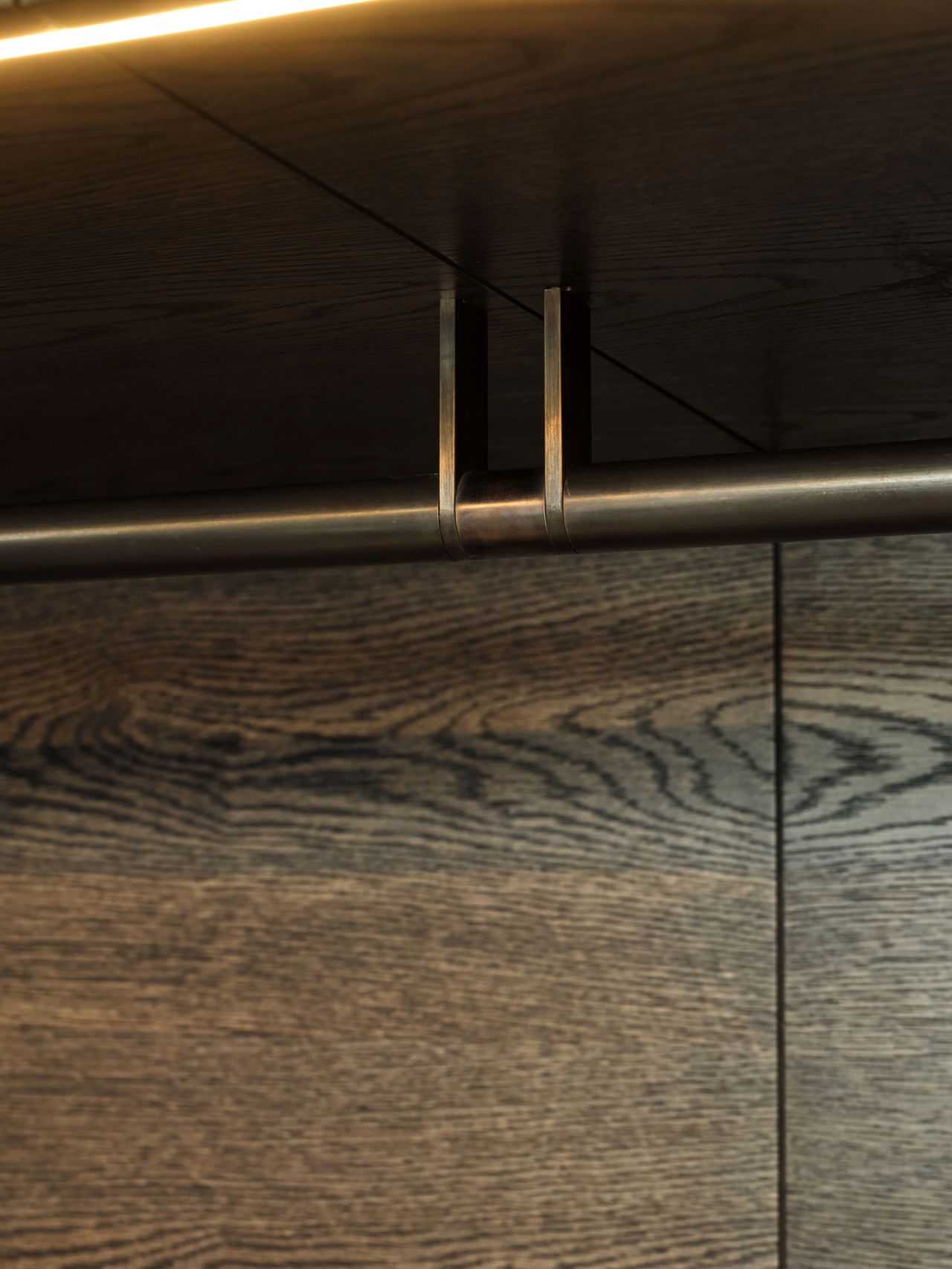 Detail of custom dark timber wardrobe cabinetry with a bronze hanging rail and integrated lighting.
