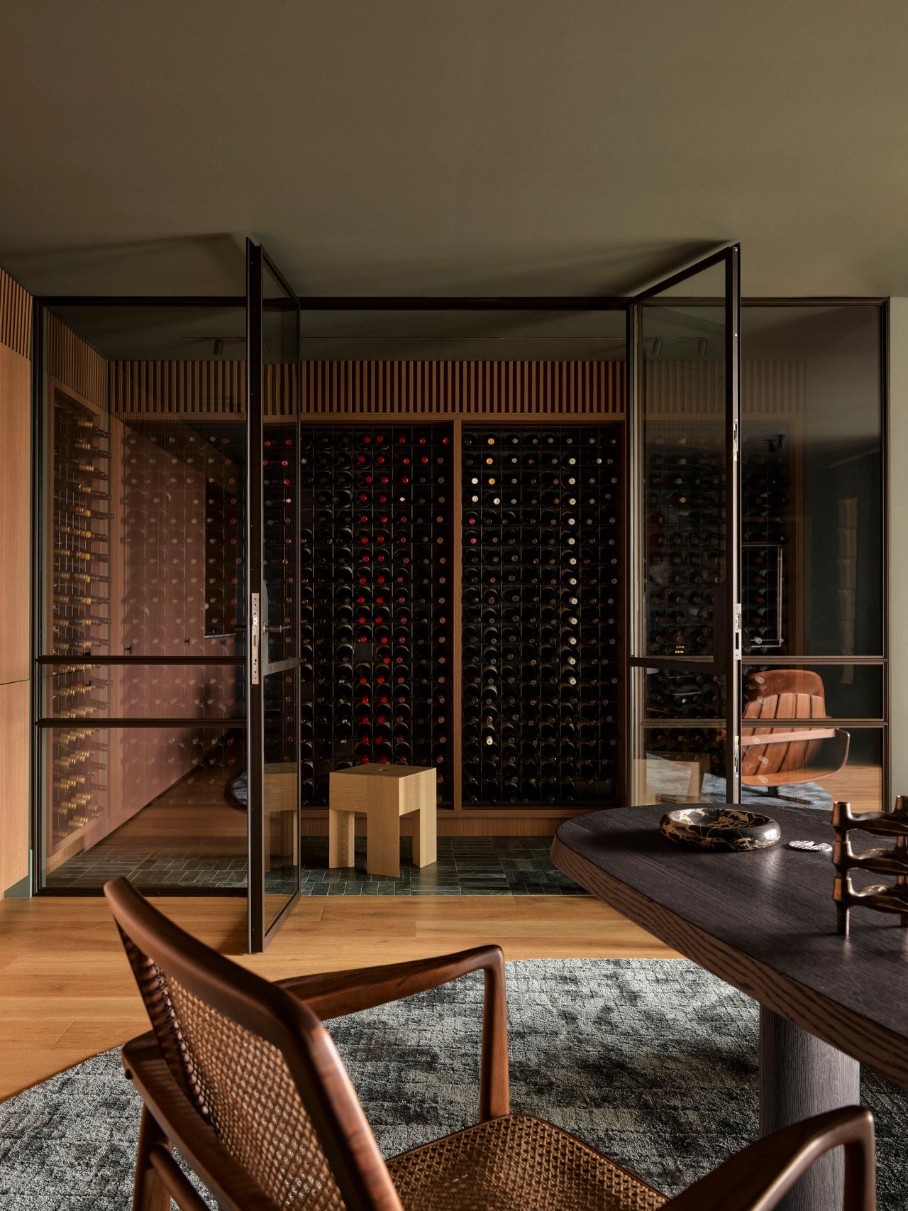 Sunset Glass-enclosed wine cellar with black steel framing and floor-to-ceiling wooden wine racks, viewed from a dining area with a dark oval table and woven-back chairs