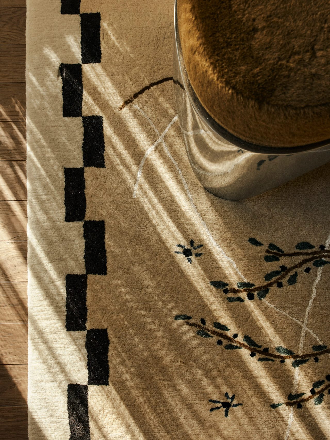Queenie Close-up of a patterned rug with a black-and-white checkered border and embroidered branch and star motifs, partially shadowed by dappled sunlight, with a brown boucle ottoman edge visible.