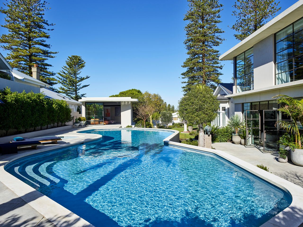 A sculptural swimming pool surrounded by lush landscaping and mature pine trees, connecting the heritage coastal residence with its contemporary extension.