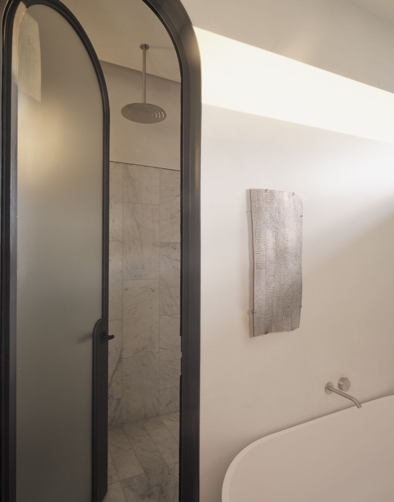 Monsieur G Bathroom with an arched black-framed shower enclosure and marble-tiled shower featuring a ceiling-mounted rain showerhead, a sculptural silver wall sconce beside a freestanding bathtub, and a wall-mounted tub spout