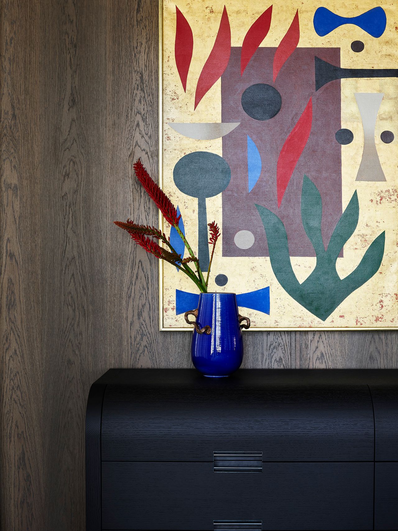 A vibrant cobalt vase and sculptural red flowers echo the bold colours of an abstract artwork, contrasting with the dark timber joinery in the contemporary interior.
