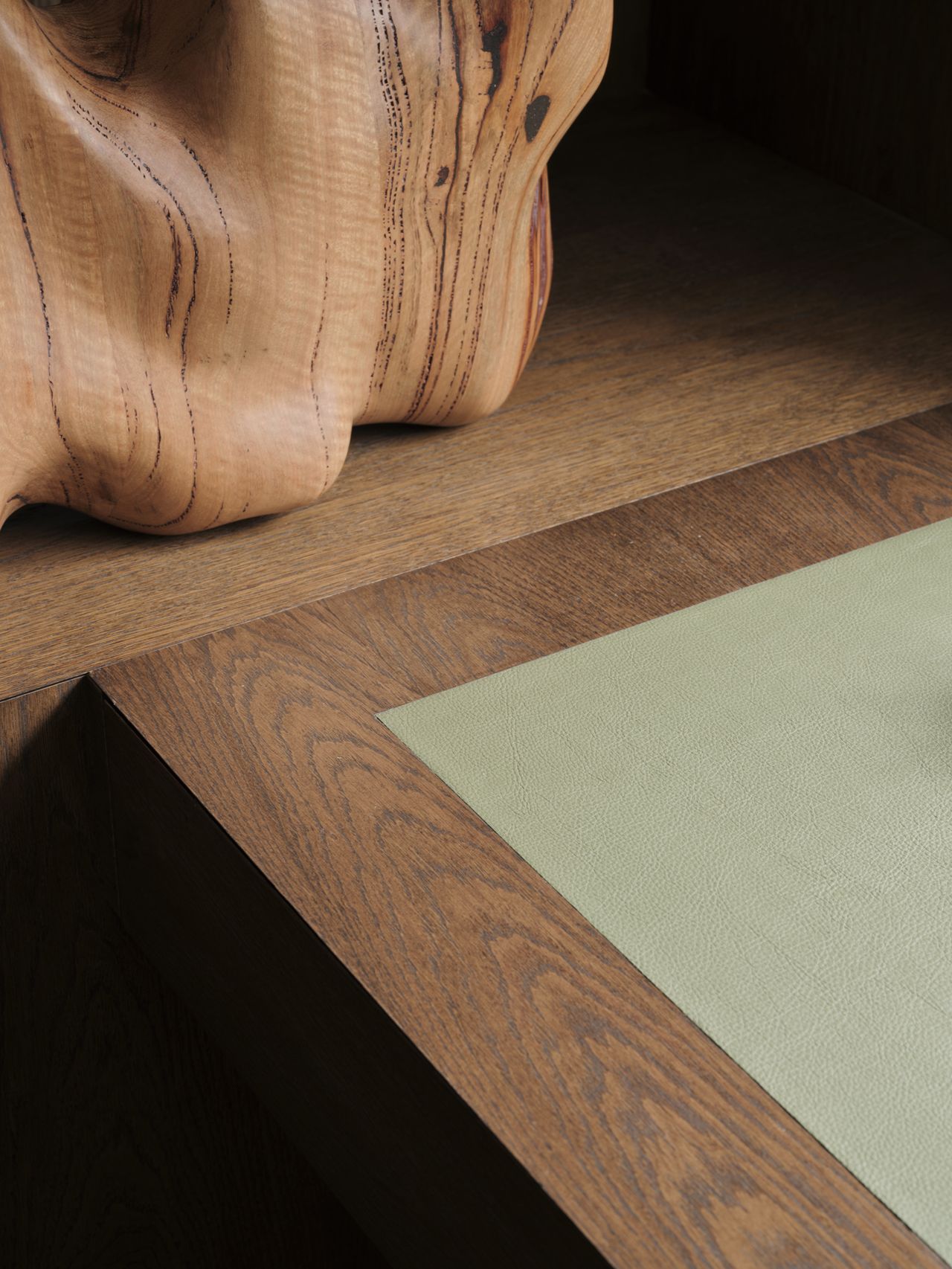 Monsieur G Detail shot of a timber desk corner with a green leather inset writing surface, showing the wood grain joinery beside a carved organic wood sculpture on the shelf above