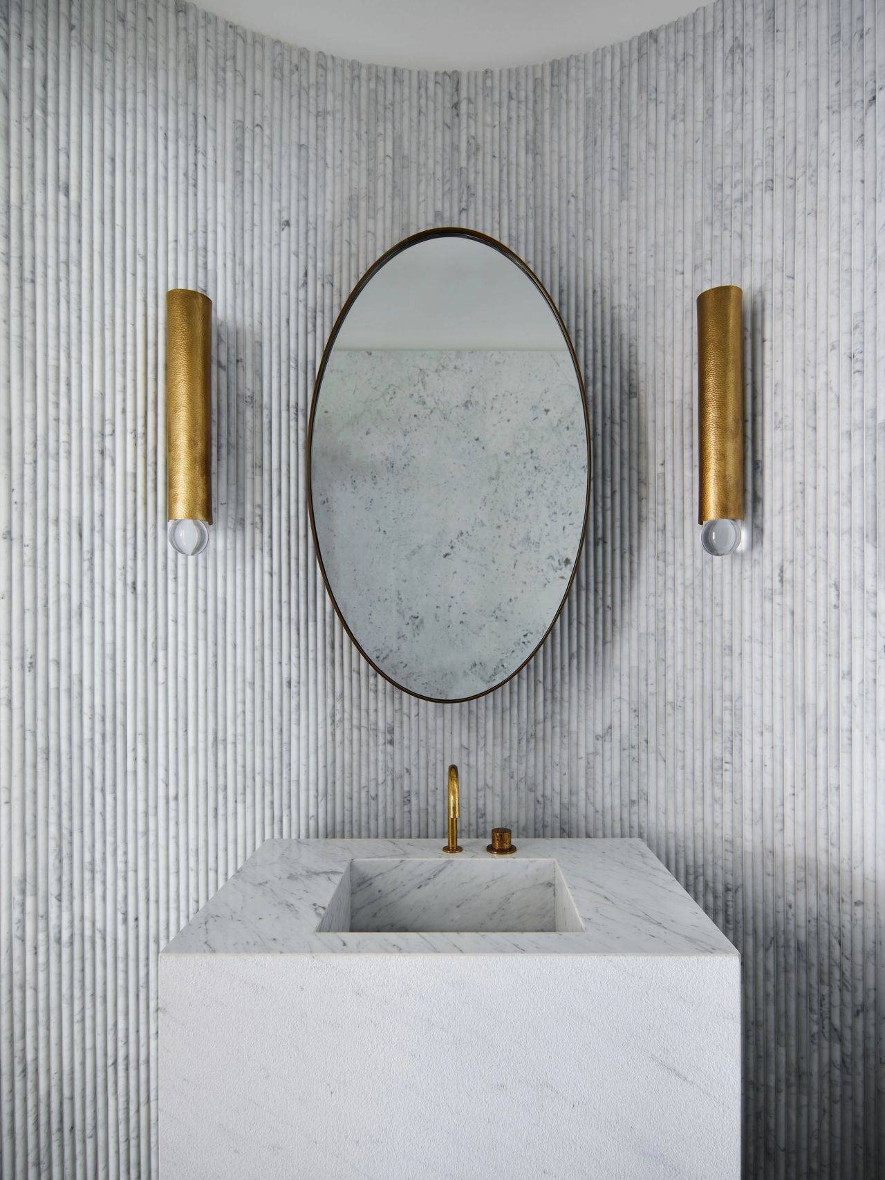 Sculptural powder room with fluted white marble walls, a monolithic marble vanity, oval mirror and symmetrical brass wall sconces.