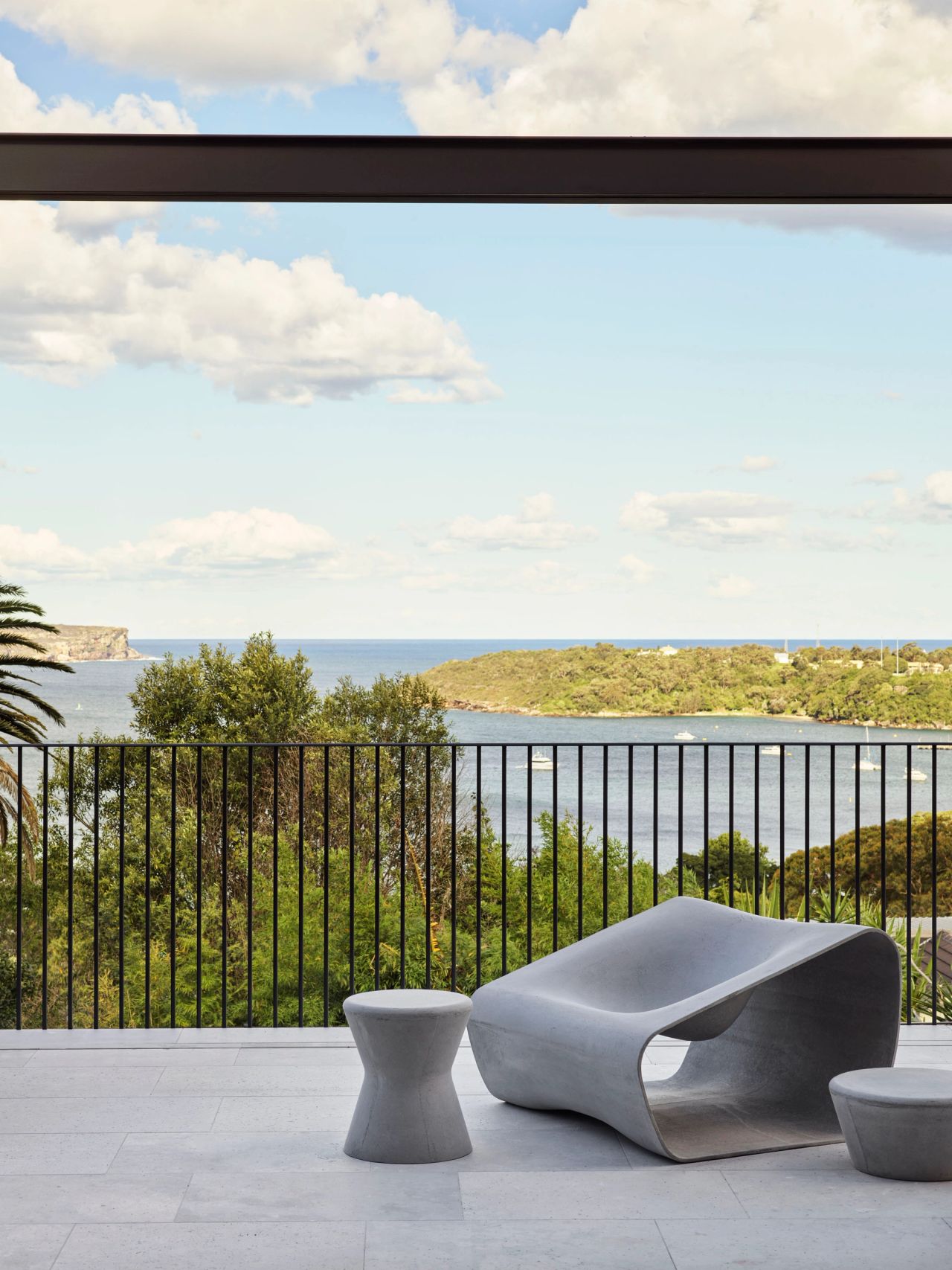 Ultramarine Sculptural grey stone lounge chair with matching side tables on a terrace overlooking a coastal harbour with boats and green headlands beyond a black railing