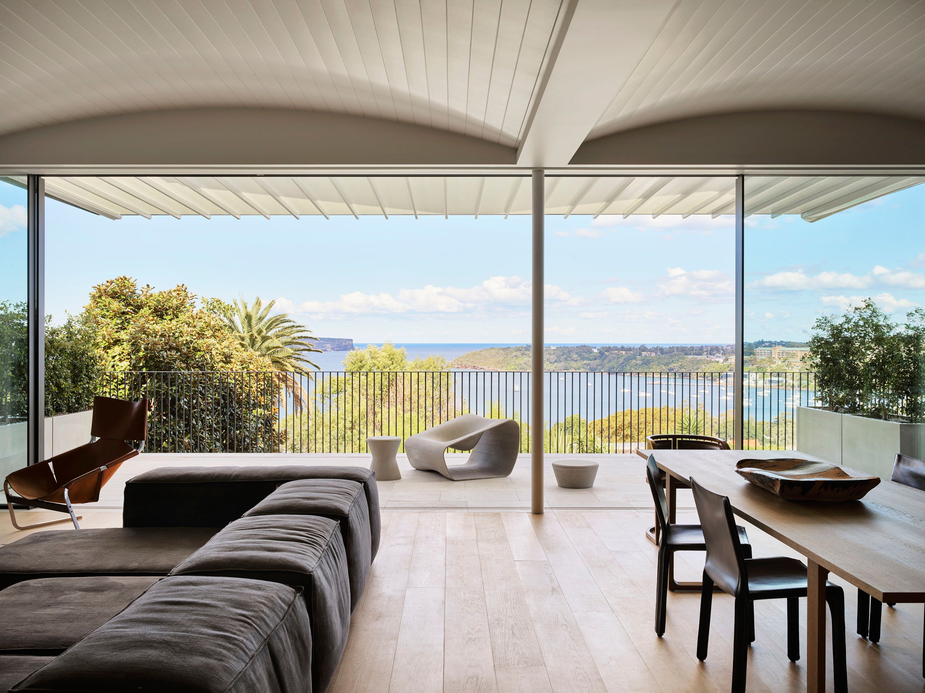 Ultramarine Open-plan living space with a charcoal sectional sofa and leather lounge chair opening onto a terrace with a sculptural stone chair, framed by a sweeping coastal harbour view
