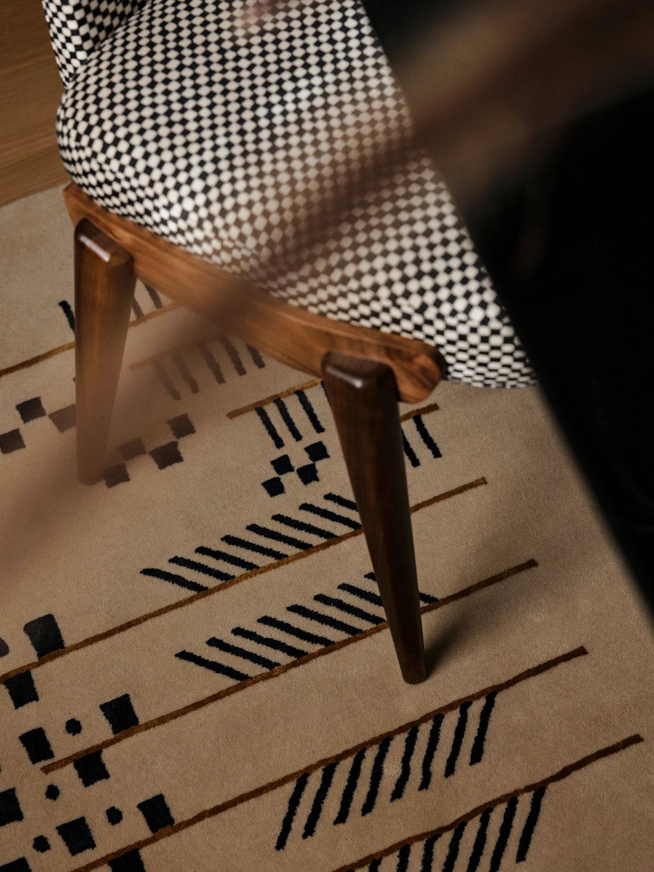 Queenie Close-up of a dark wood chair leg with houndstooth-patterned upholstery on a graphic cream rug featuring black stripes, dashes, and checkered motifs