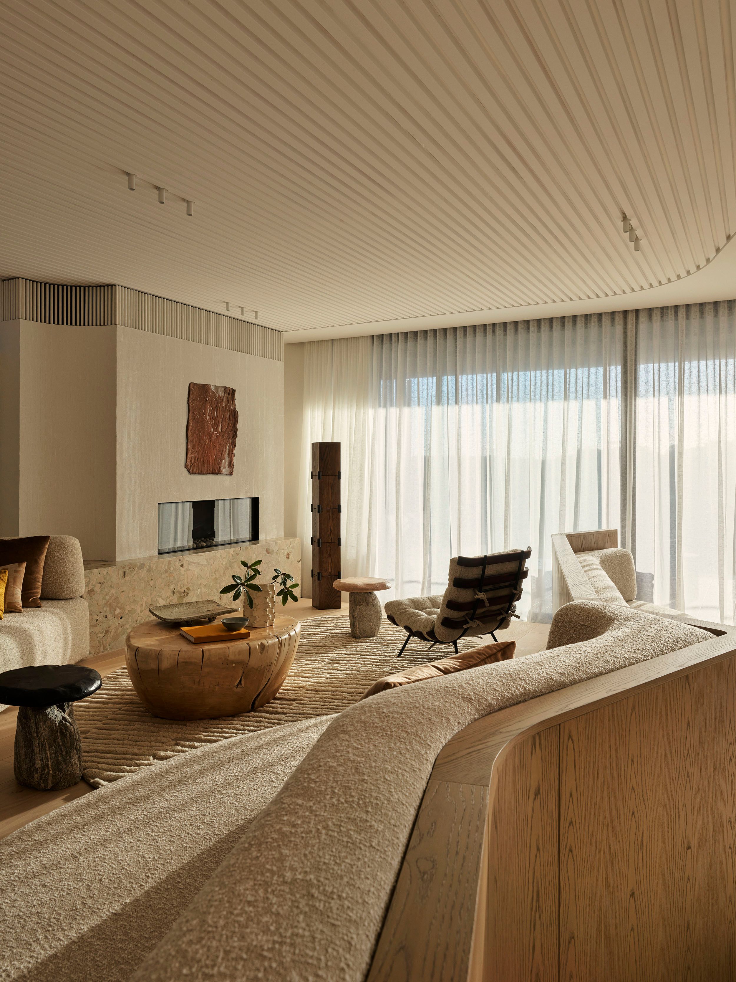 Patchwork Neutral-toned living room with a curved boucle sofa, sculptural wood stump coffee table, mushroom-shaped stone side table, black leather-strap armchair, and sheer floor-to-ceiling curtains, with a rustic textured artwork above a built-in fireplace.