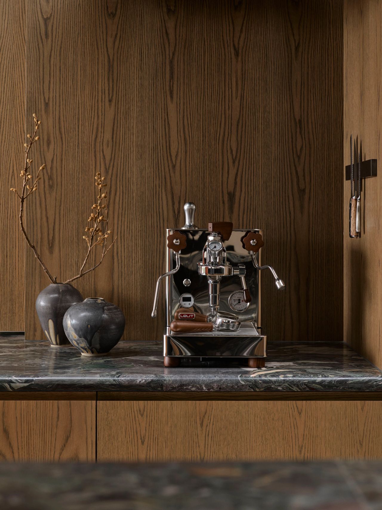 Patchwork Chrome espresso machine with tan leather-wrapped handles on a dark marble countertop, flanked by two grey ceramic vases with dried branches, set against warm oak-panelled cabinetry.