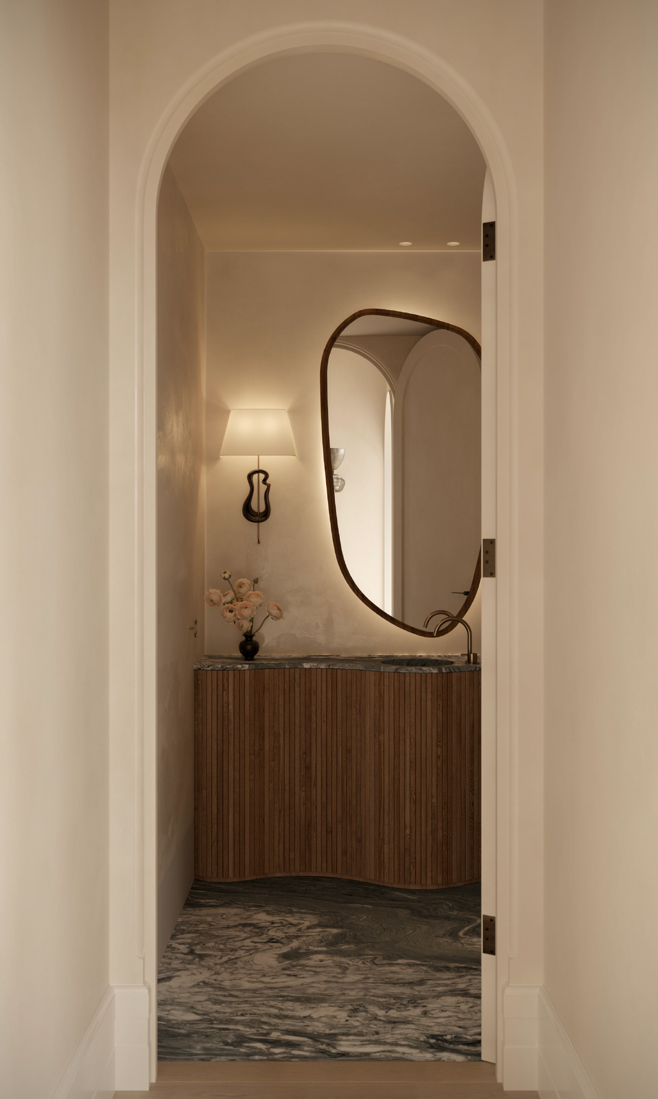Lady M View through an arched doorway into a powder room with a curved fluted timber vanity, dark stone countertop and floor, an organic-shaped brass-framed mirror, and a small table lamp beside a sculptural wall sconce