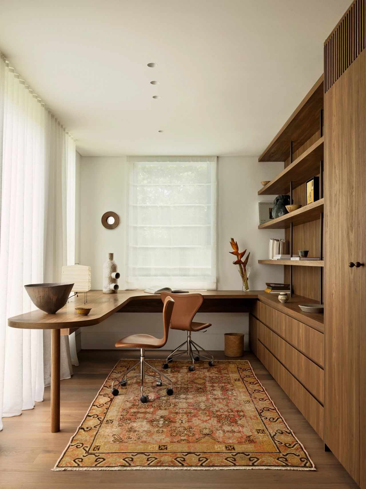 Dancing On The Ceiling Home office with a curved wooden desk, tan leather swivel chair, built-in wood shelving styled with books and ceramics, and a patterned vintage-style rug, softly lit by sheer curtains