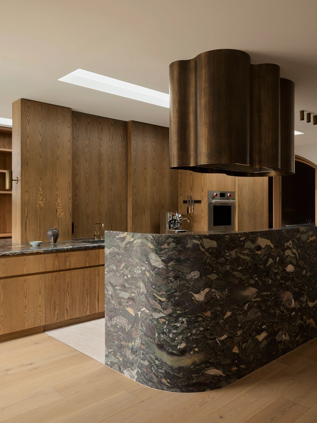 Patchwork Sculptural kitchen with a curved, dark breccia marble island base featuring dramatic cream, tan, and grey veining, paired with warm oak cabinetry and a fluted bronze range hood in an organic wave shape suspended above the cooktop.