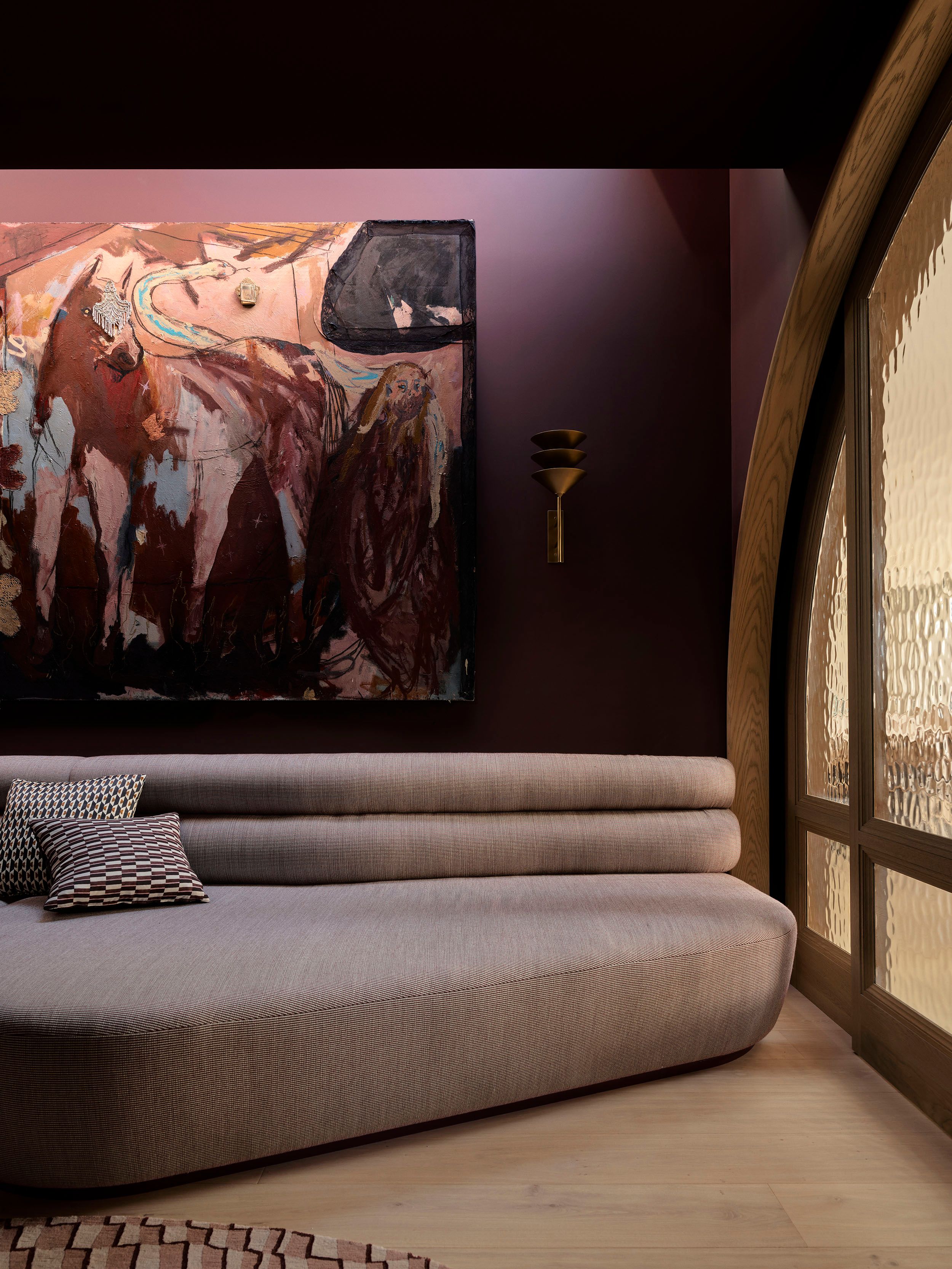 Patchwork Media Room Moody, deep-plum living room with a large abstract expressionist painting in warm earth tones, a low ridged sofa with patterned cushions, a brass wall sconce, and an arched wooden-framed window with textured glass.