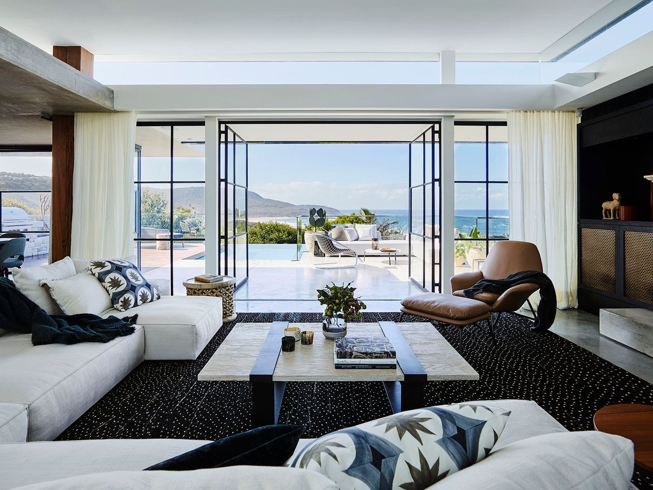 Killcare Open-plan living room with white sectional sofas and patterned cushions around a stone coffee table on a dark textured rug, black steel-framed doors opening to a pool terrace with sweeping ocean and headland views, and a dark timber cabinet with decorative objects to the side
