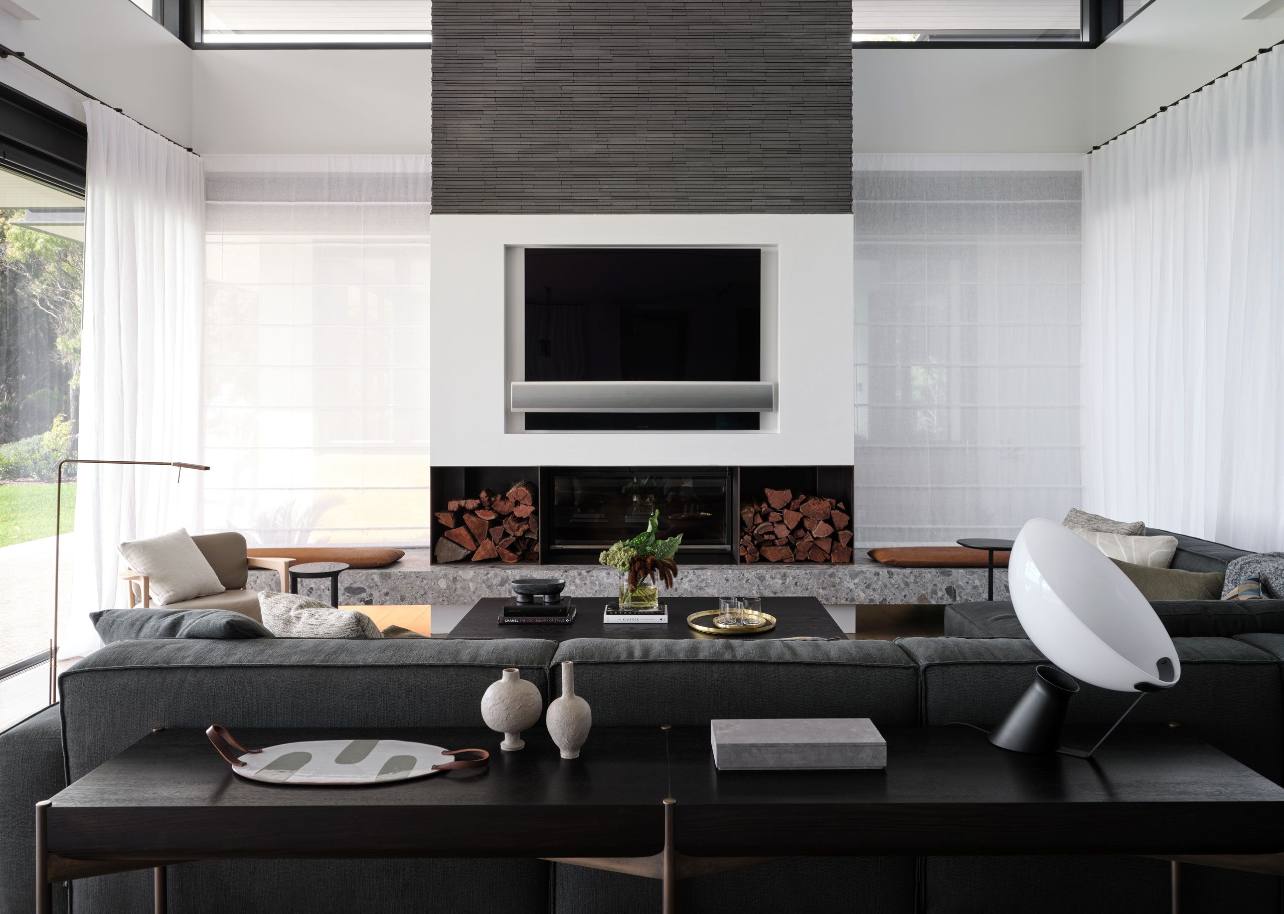 Contemporary living room with charcoal sectional sofa, central fireplace, built-in log storage and sheer white curtains.