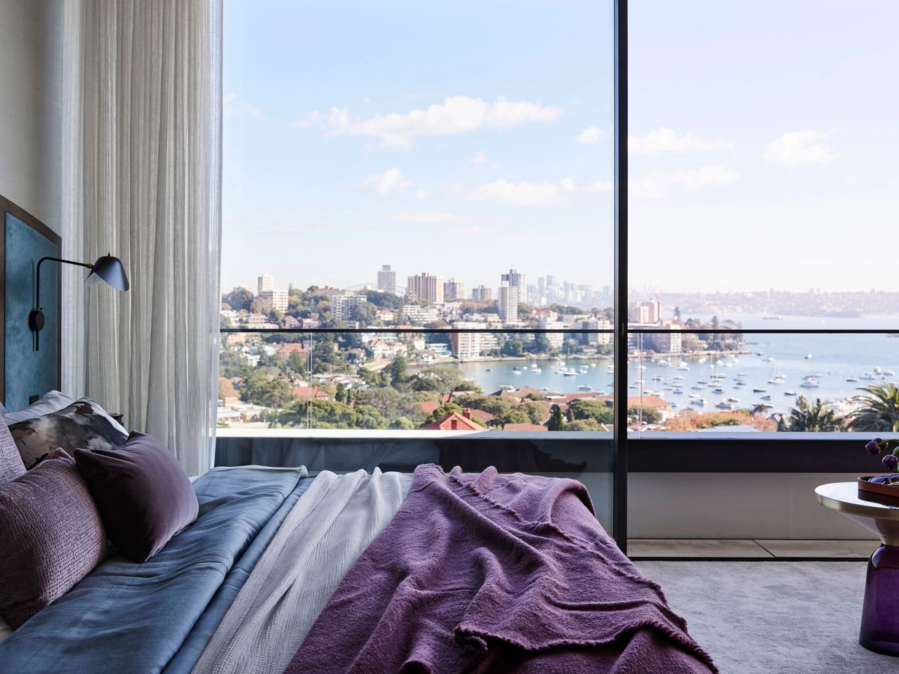 Luxury bedroom with floor-to-ceiling windows, layered purple and blue bedding, and panoramic harbour and city views.