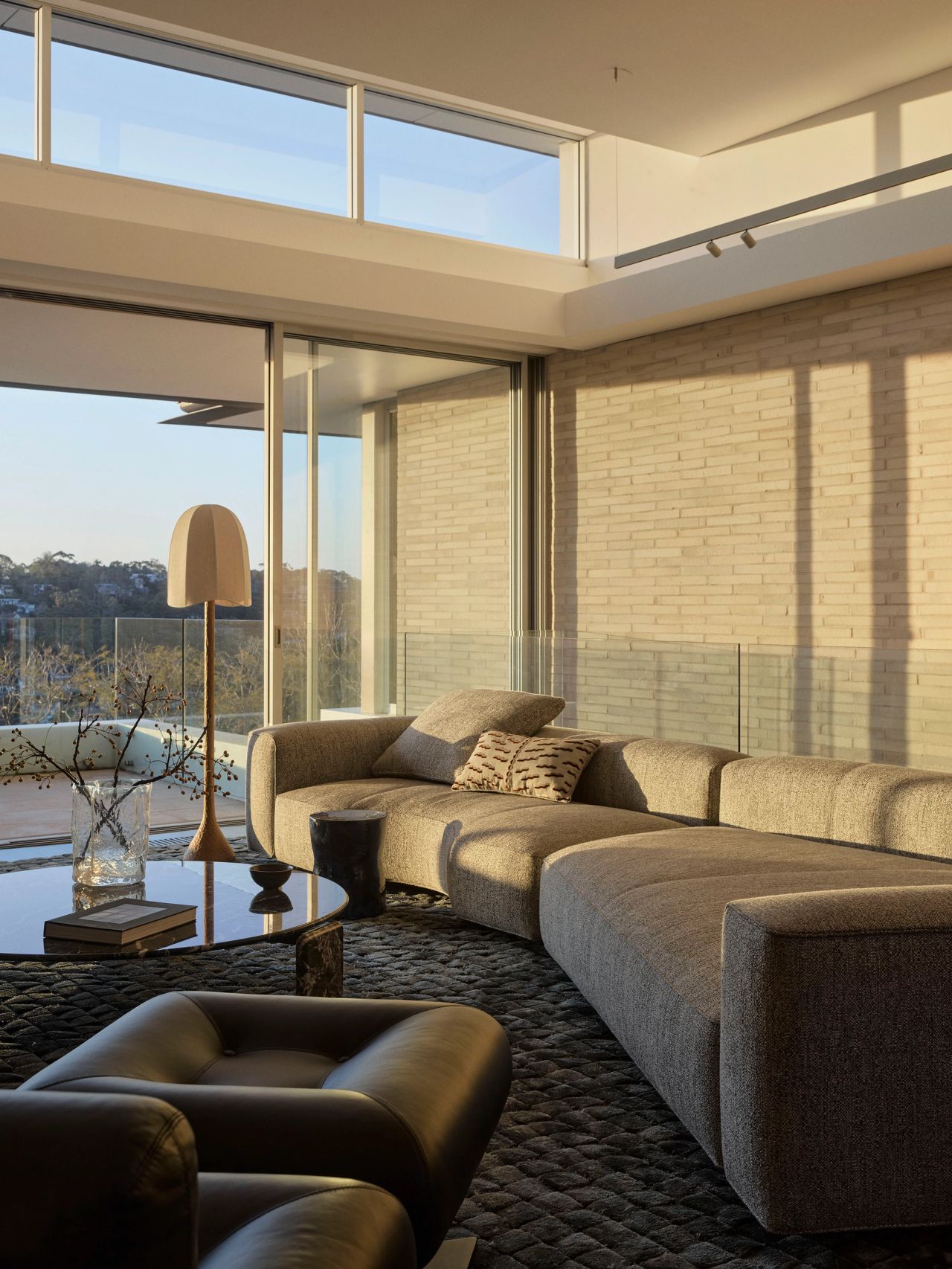 Sunset Sunlit living room with a curved beige sectional sofa, black leather ottoman, mushroom-shaped table lamp, and floor-to-ceiling glass doors opening onto a terrace with treetop views