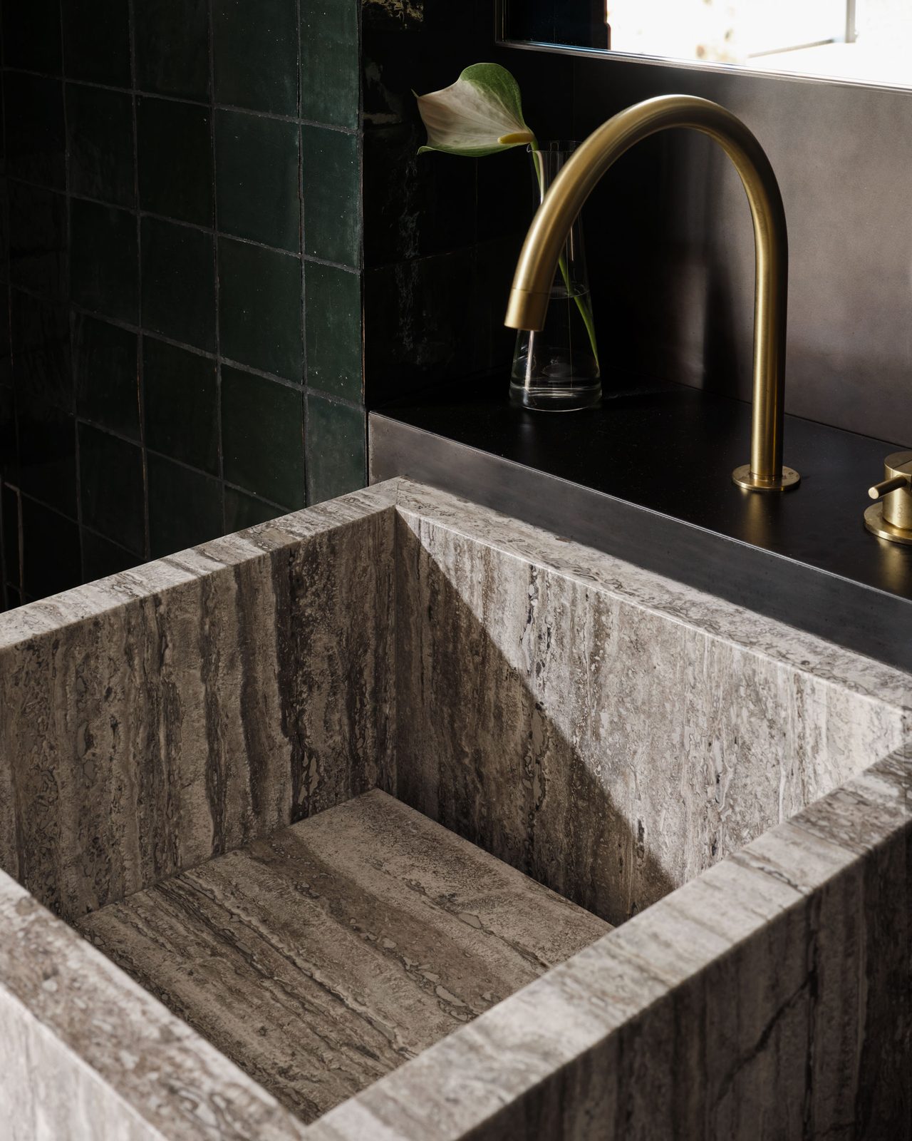 Close-up of a sculptural grey stone basin with curved brass tapware and deep green tiled walls.