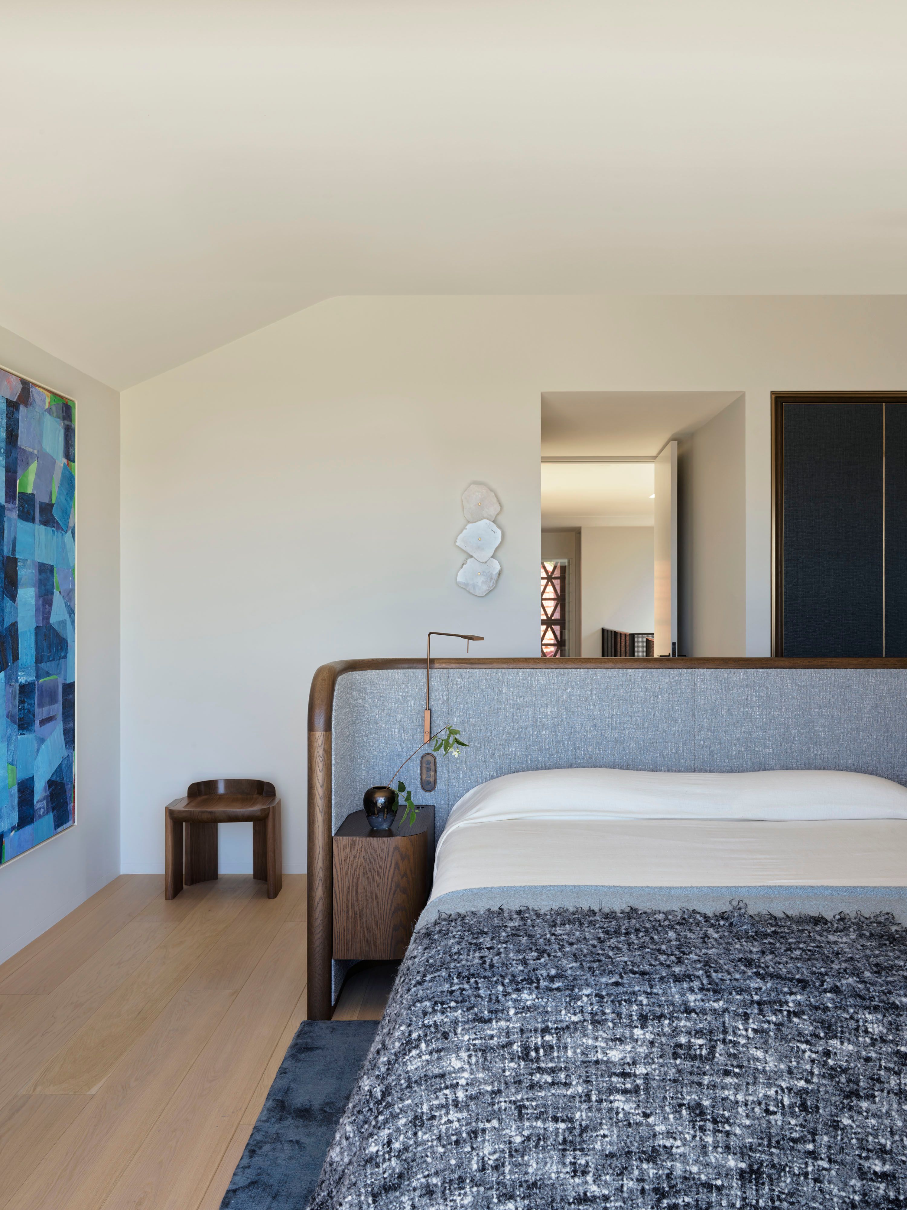 Ultramarine Bedroom with a curved wood-framed grey upholstered headboard, textured charcoal throw, a sculptural wooden stool, and a wall-mounted disc sculpture, opening onto a hallway beyond.