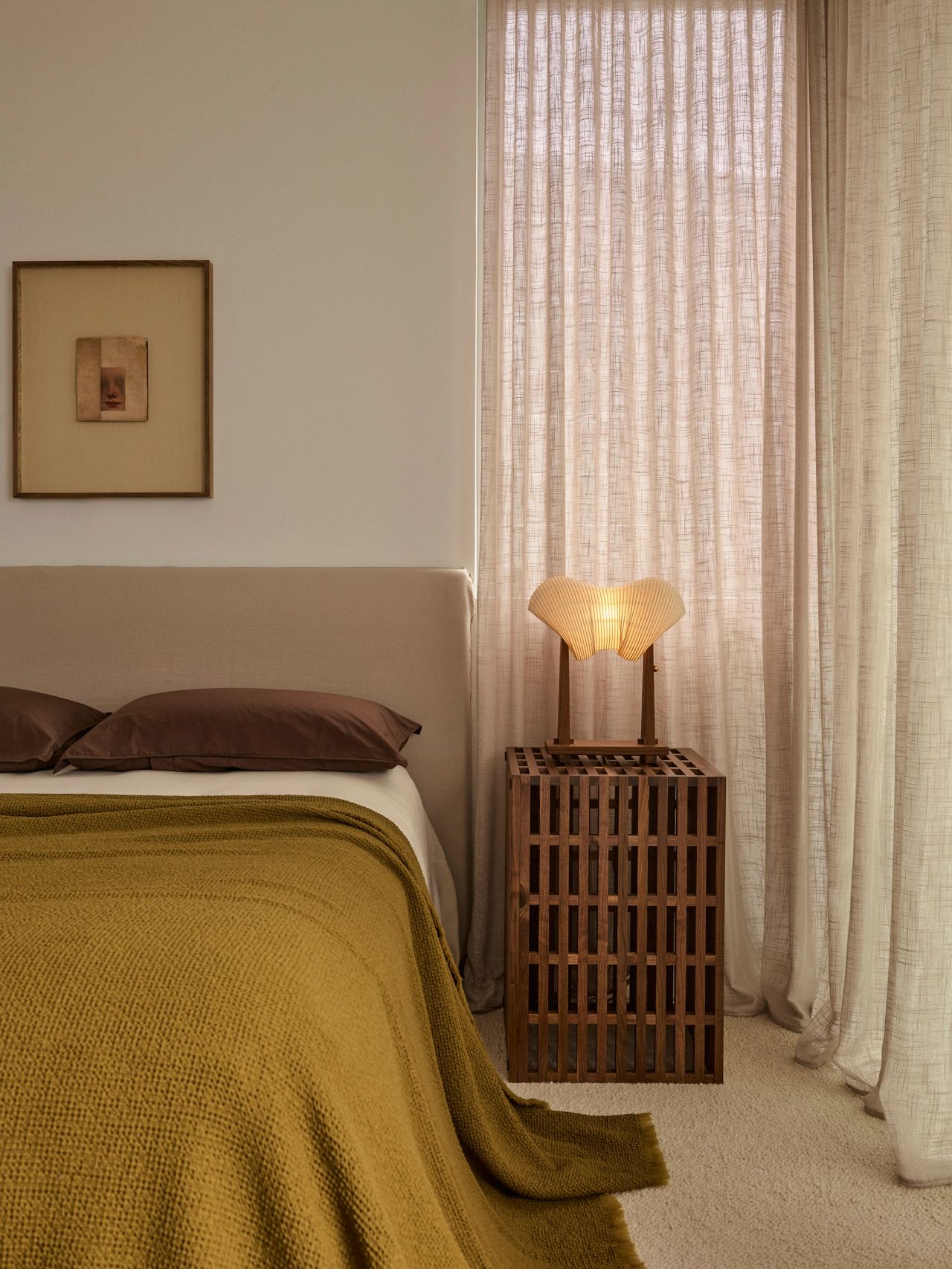 Orla contemporary bedroom blending Brazilian influences, with a beige upholstered bedhead, brown pillowcases, and a mustard throw at the foot of the bed. A walnut lattice-detail side table holds a paper lamp-style bedside light, set against heavy linen drapes and cream carpet.