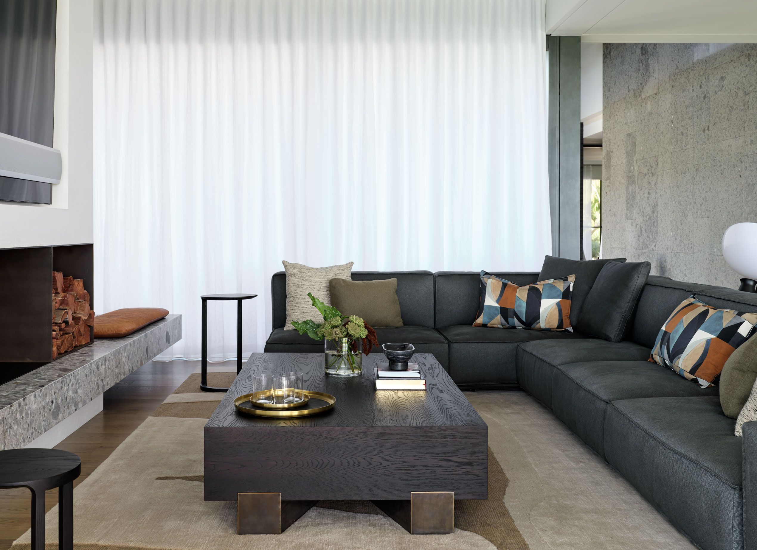 Sophisticated contemporary living room featuring a deep charcoal sectional, graphic cushions, sheer curtains and a sculptural dark timber coffee table.