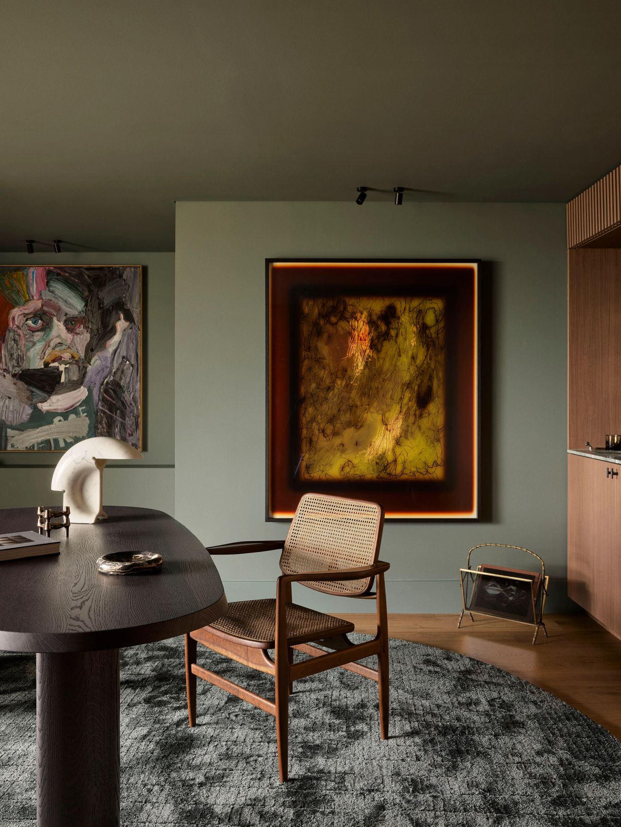 Sunset Sage green room with a moody abstract painting glowing beneath a spotlight, a cane-back dining chair, a dark oval table, and a colourful figurative artwork on the adjacent wall