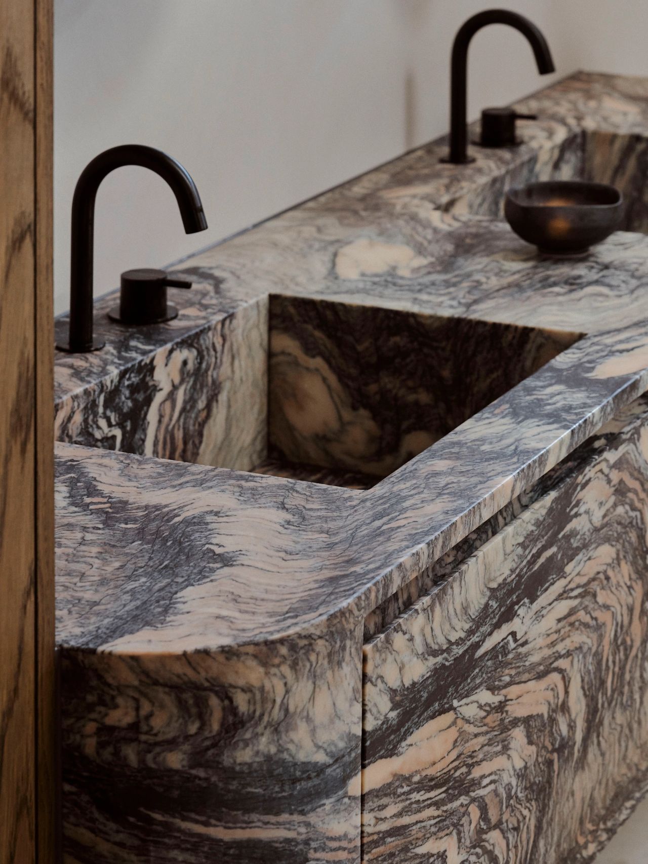 Patchwork Sculptural double vanity carved from dramatic swirling grey and beige marble, with an integrated basin, black gooseneck tapware, and a small black ceramic bowl on the counter.