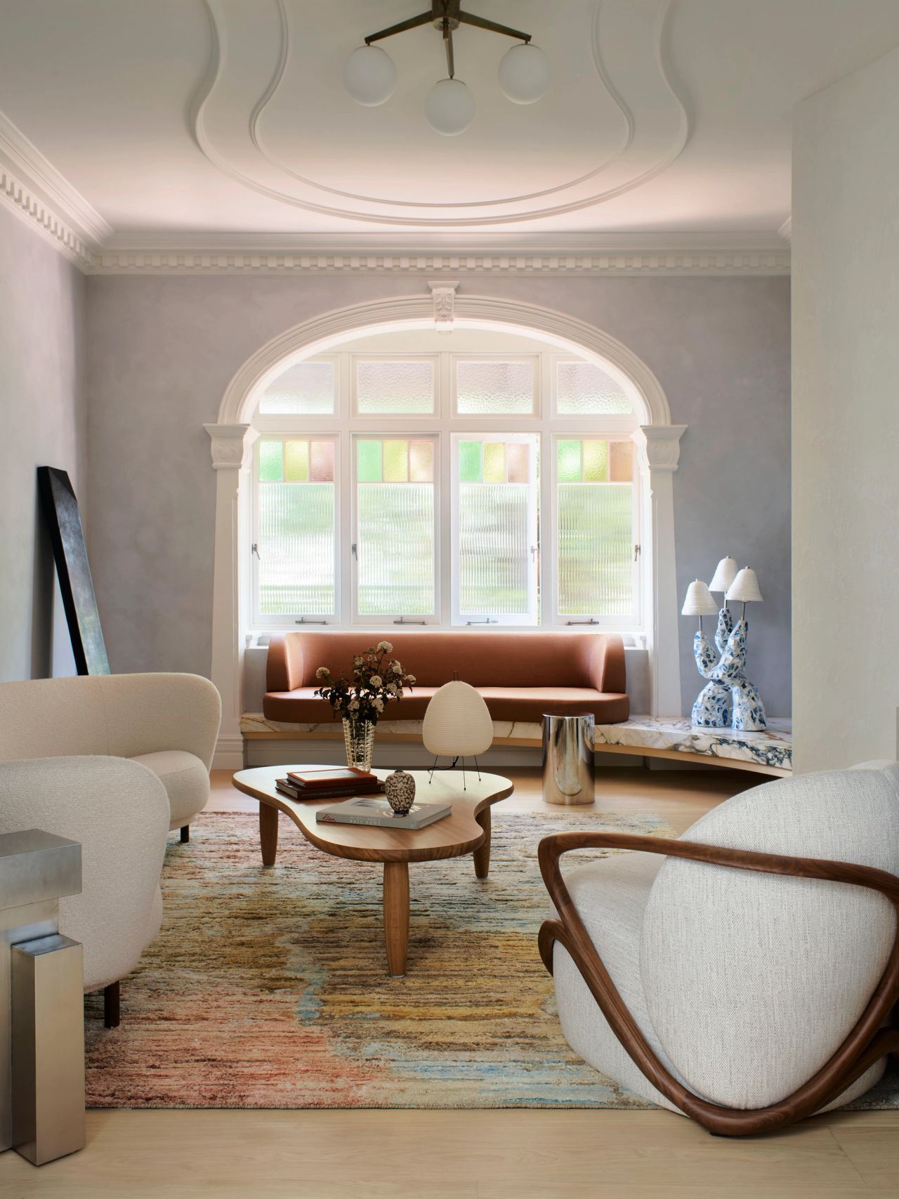 Ultramarine Sitting room with an arched period window featuring stained glass panels above a built-in terracotta banquette, a curved white bouclé armchair, an organic-shaped coffee table, and a decorative blue-and-white ceramic table lamp