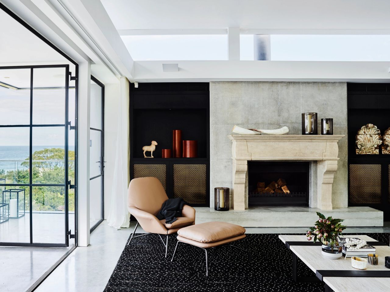 Killcare Living room with an ornate carved stone fireplace flanked by black joinery with brass mesh cabinet doors and decorative objects, a tan leather lounge chair and ottoman, and black-framed glass doors opening to an ocean view