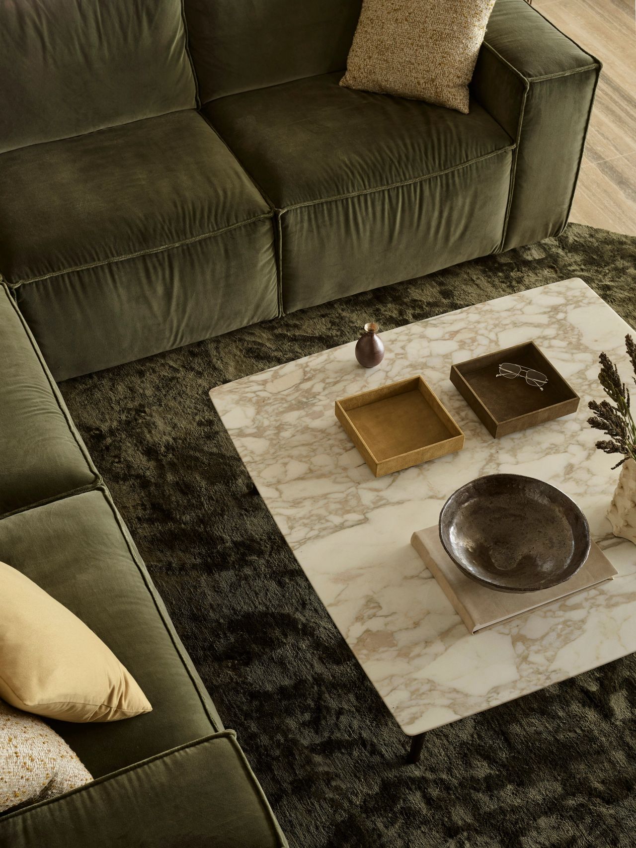 Orla contemporary living room blending Brazilian influences, birds-eye detail of a marble coffee table with suede trays in mustard and dark olive green, a forest-green velvet sofa on a forest-green silk rug