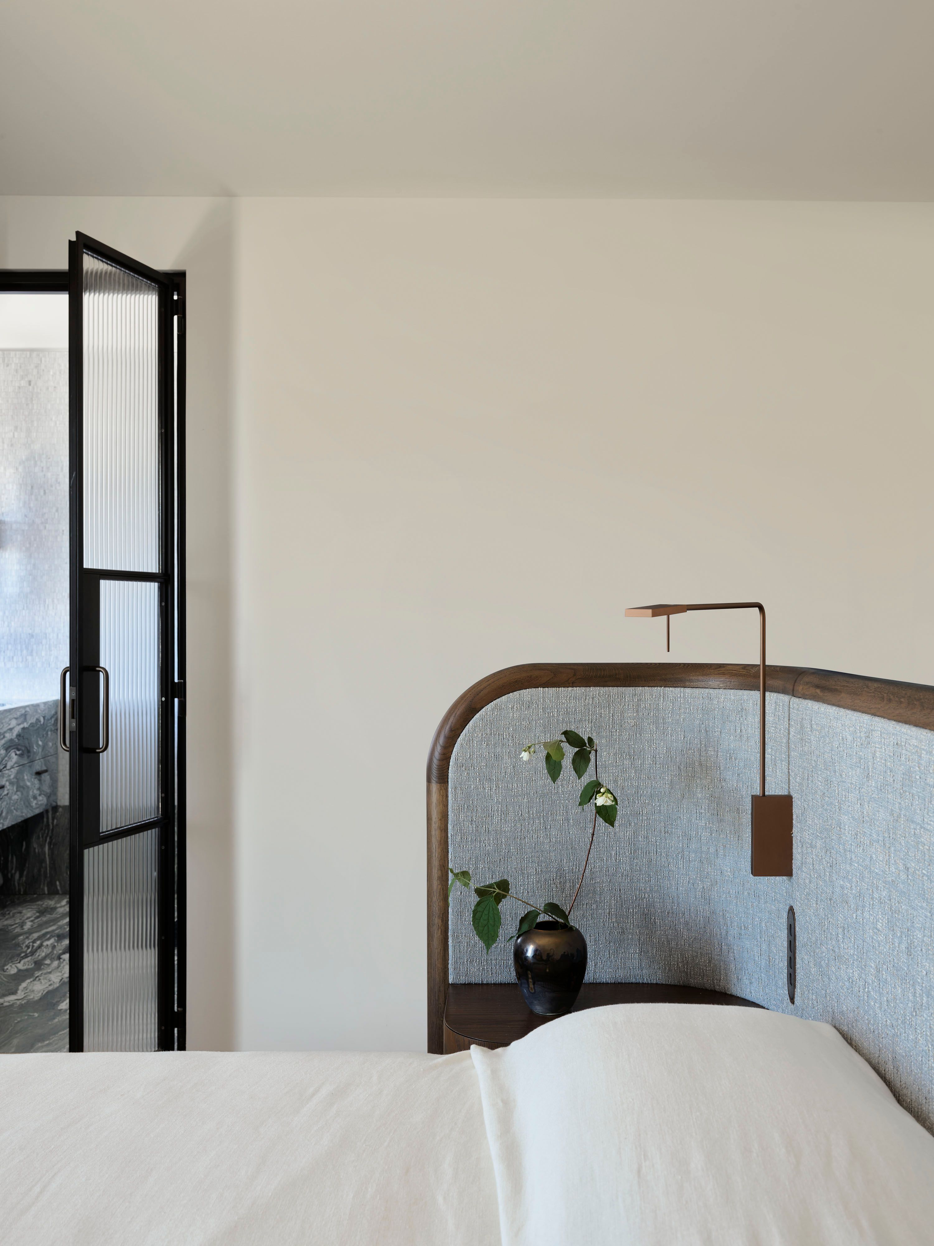 Ultramarine Minimal bedroom corner with a curved, upholstered wooden headboard, a sleek angular reading light, and a small dark vase with a leafy branch, beside a black-framed reeded glass door.