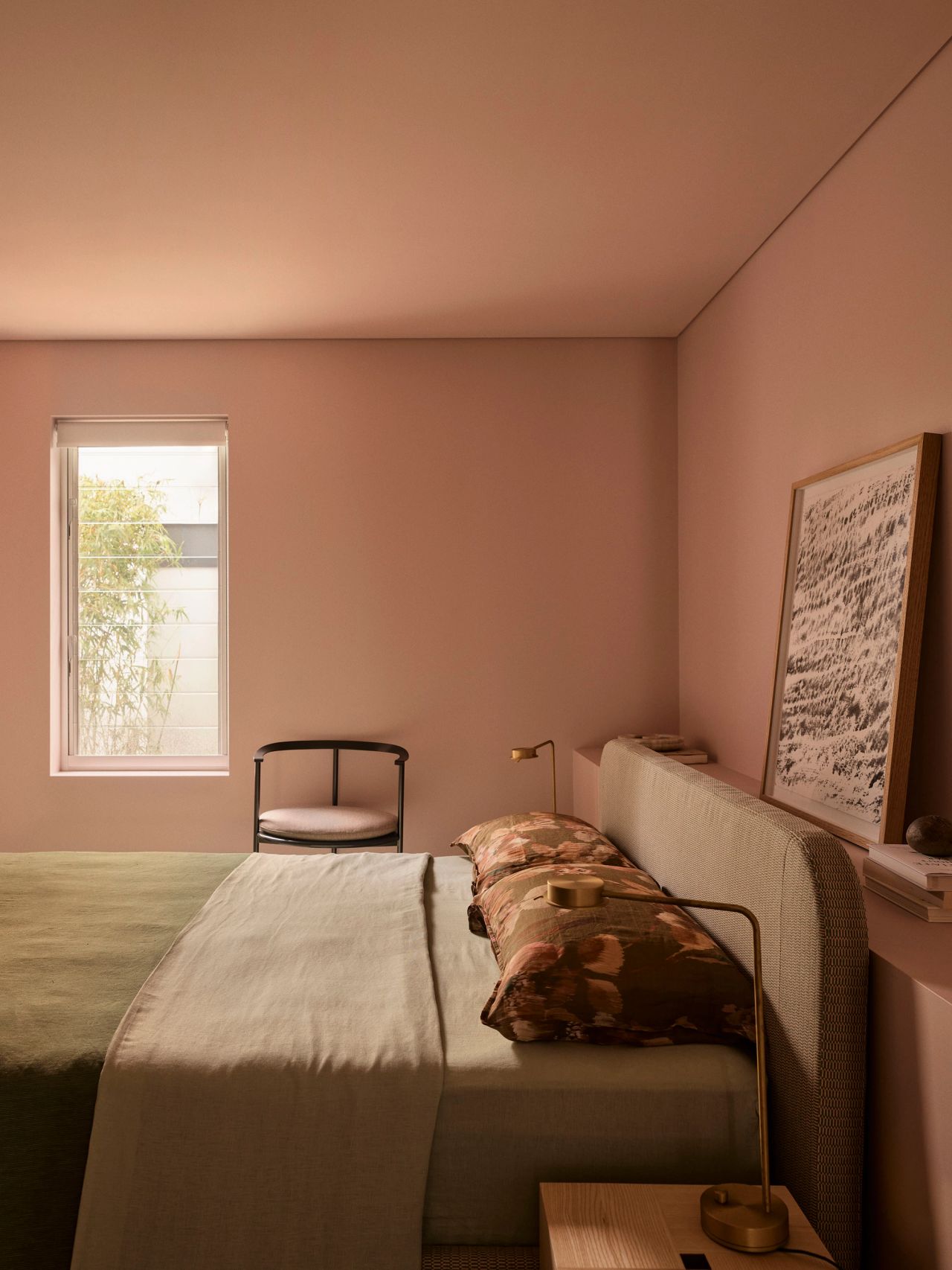 Sunset Soft pink-walled bedroom with an olive green bed, curved upholstered headboard, floral throw pillows, a brass reading light, a black wire-frame accent chair, and a black-and-white framed artwork