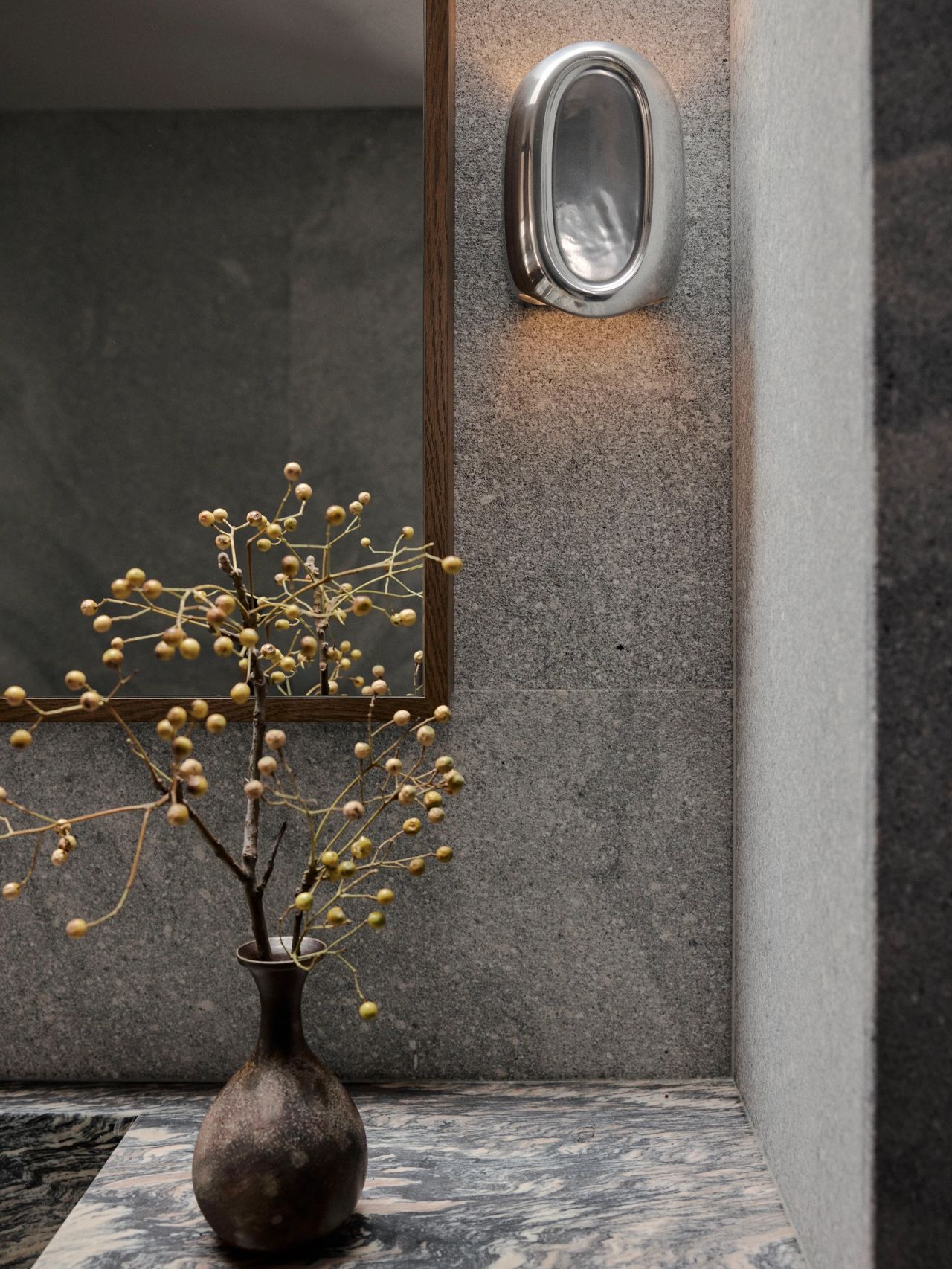 Sunset Small dark ceramic vase with berry-laden branches on a marble ledge, beside a wood-framed mirror and an oval brushed-metal wall sconce against textured stone walls