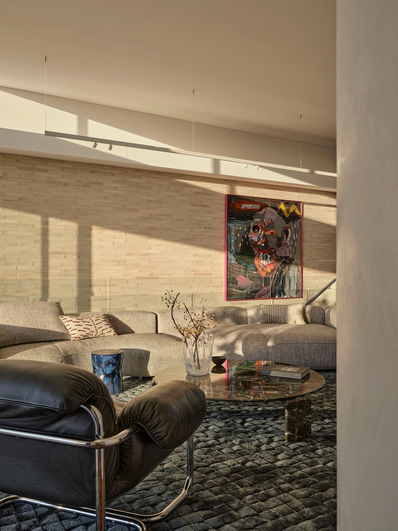 Sunset Living room with a black leather chrome-framed lounge chair, beige sectional sofa, and a colourful abstract figurative artwork on a textured stone wall, with a round marble coffee table on a chunky woven rug