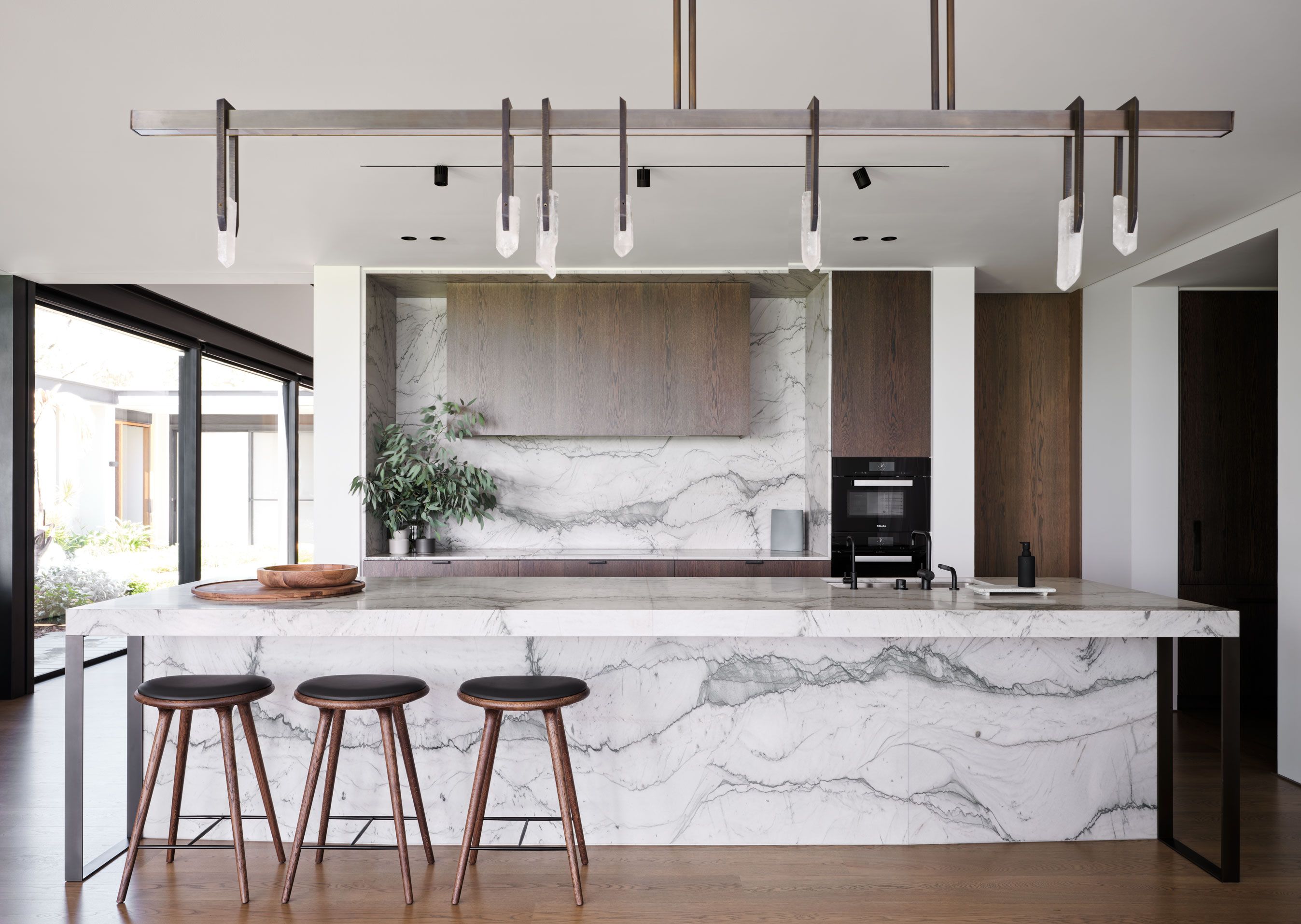 Refined contemporary kitchen featuring dramatic marble island, dark timber cabinetry, bar stools and sculptural linear pendant lighting.