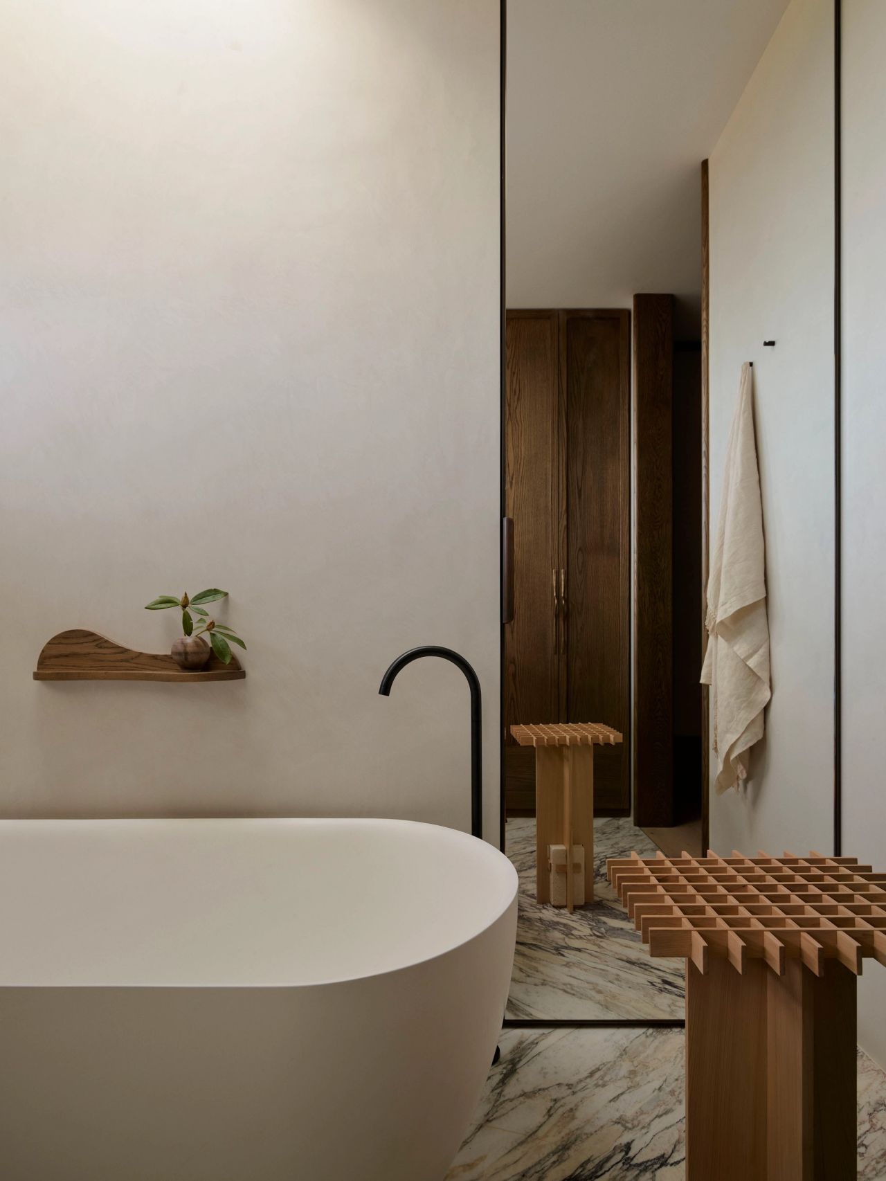 Patchwork Freestanding white bathtub with black wall-mounted tap, a sculptural wooden shelf holding a small plant, and a slatted wooden bench on marble flooring, reflected in a mirror leading to a wardrobe beyond