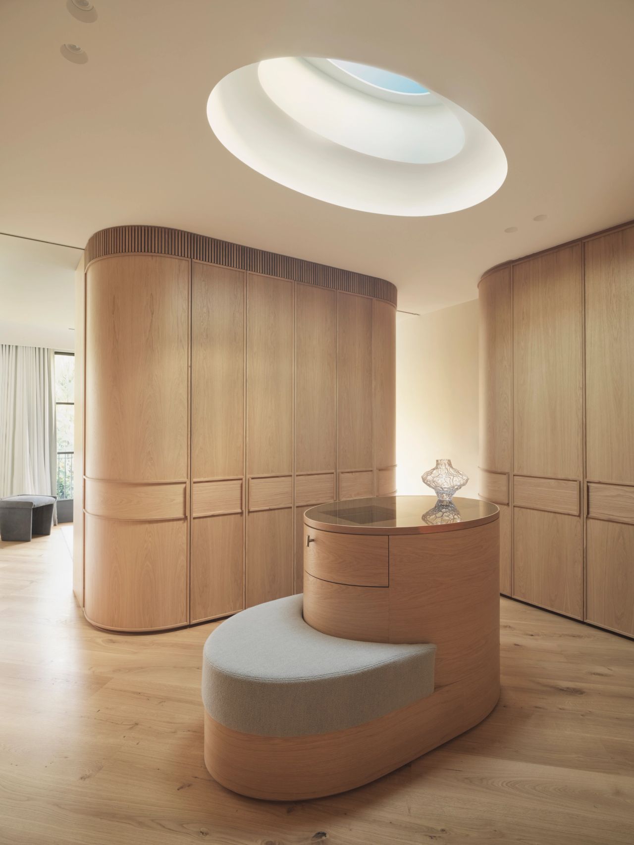Monsieur G Curved oak dressing room with a rounded oval skylight overhead, floor-to-ceiling wardrobe joinery with fluted detailing, and a curved timber dressing table with an upholstered ottoman seat and a decorative glass perfume bottle