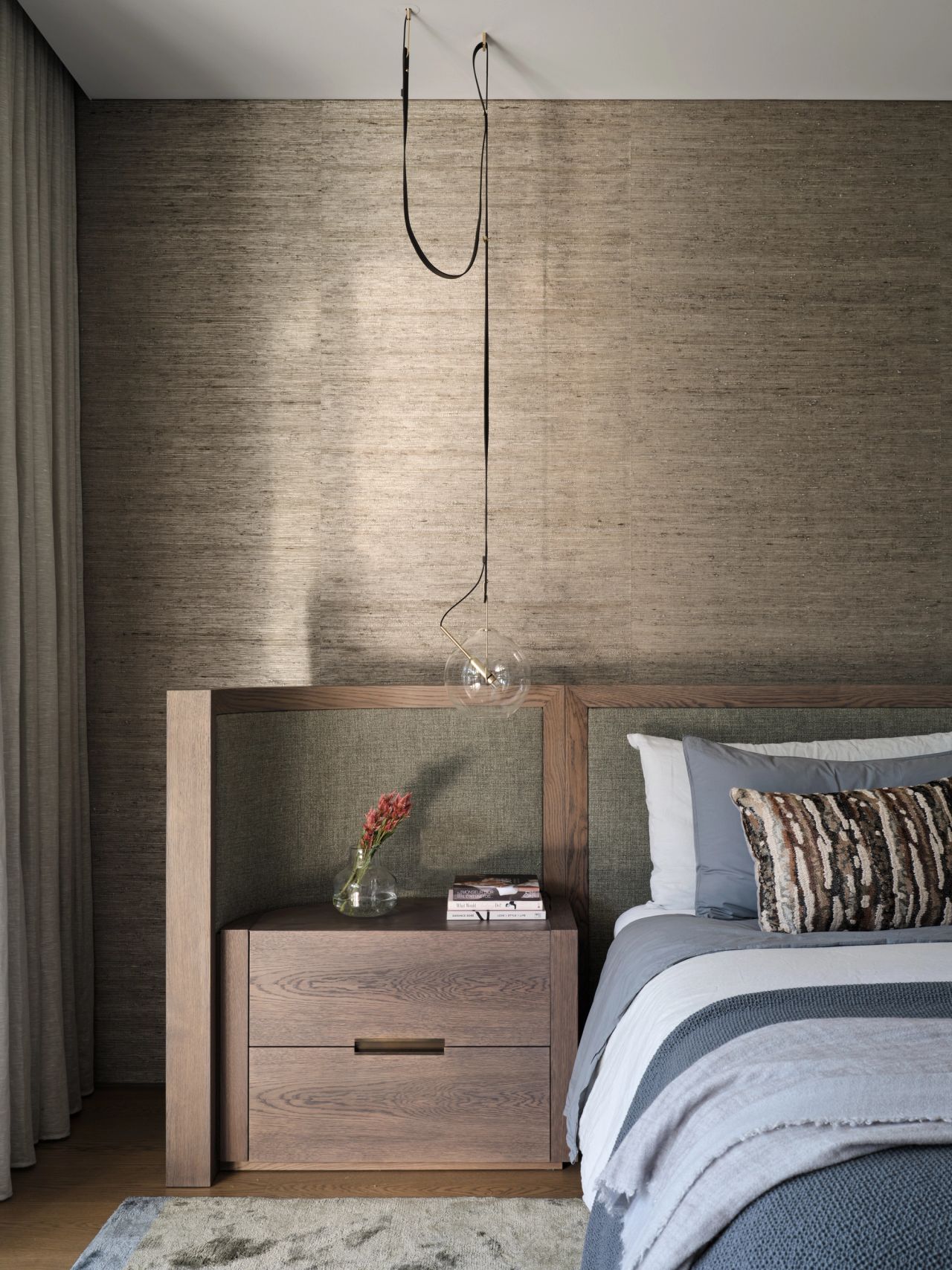 Serene contemporary bedroom layering warm timber, textured upholstery and soft grey-blue bedding beneath a sculptural pendant.