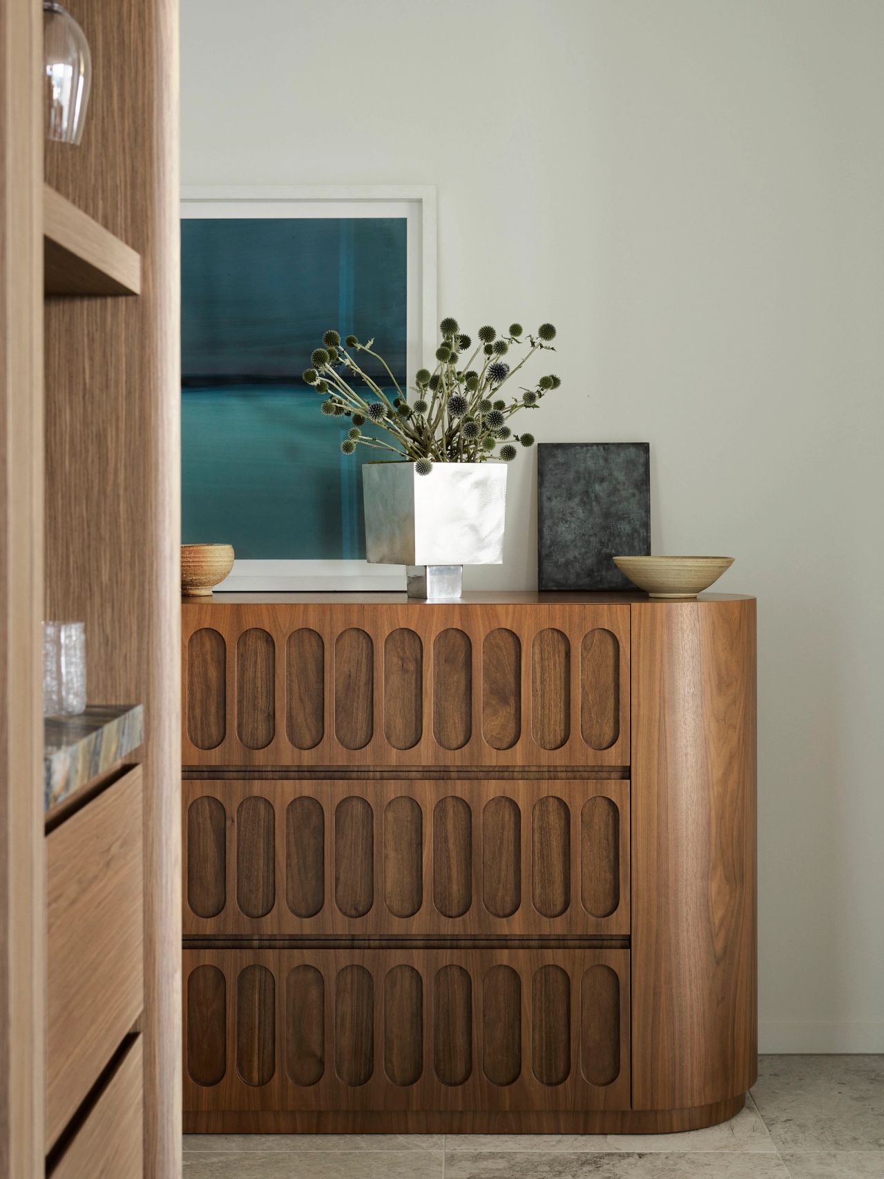 Rich timber joinery with rounded sculptural detailing, layered with ceramics, foliage, and ocean-toned artwork.