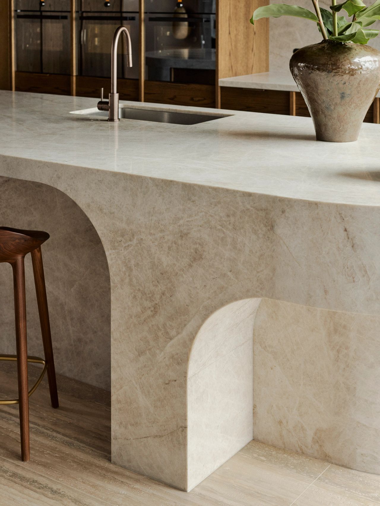 Orla contemporary kitchen island detail in white/beige quartzite granite - sweeping arch detail in base of island block, timber stool and ceramic vase on counter top